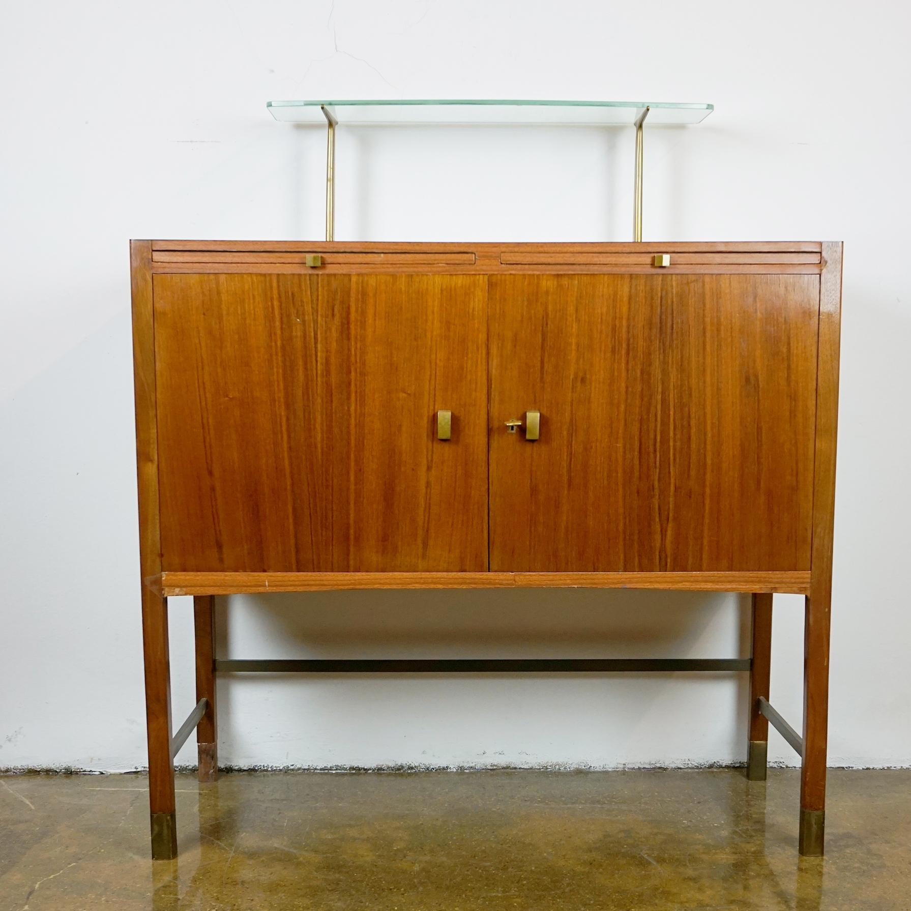 This elegant small cabinet or credenza or sideboard was designed by the Austrian architect and designer Oswald Haerdtl and manufactured in the 1950s in Vienna. 
This charming cabinet is made of Walnut and brass and impresses with its pure design