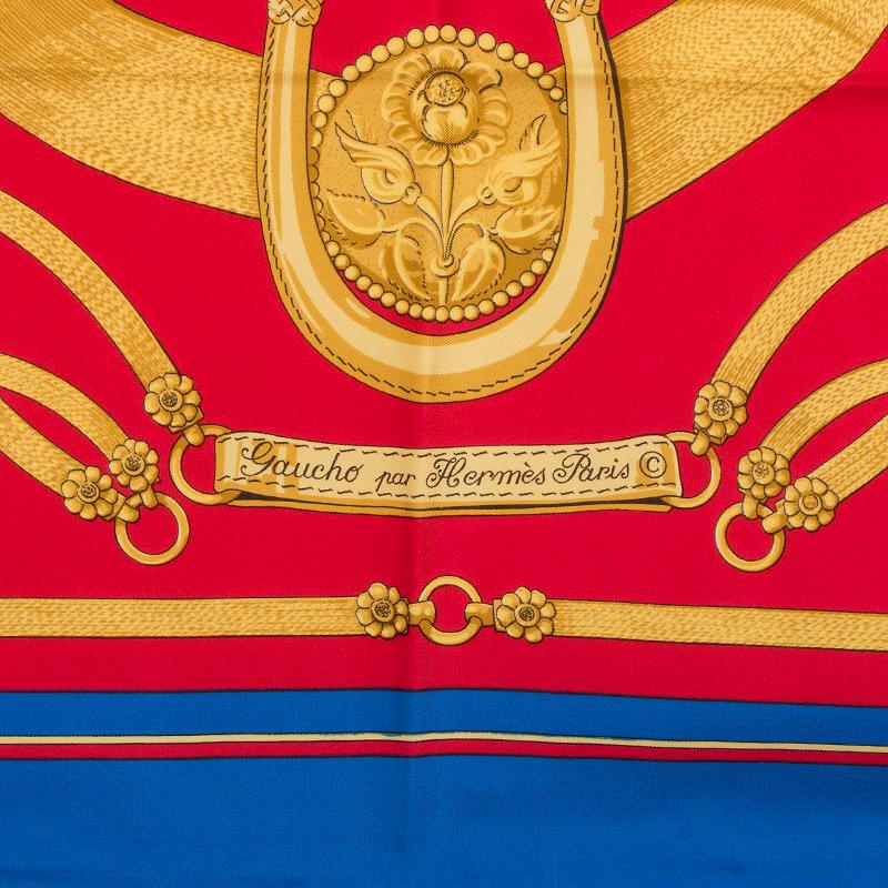 Hermes 'Gaucho 90' scarf by Cathy Latham in red silk twil (100%) with royal blue border and details in gold. Has been worn and in excellent condition. There's a printing error in the corner about the size fo a dime.

Width 90cm (35.1in)
Height 90cm