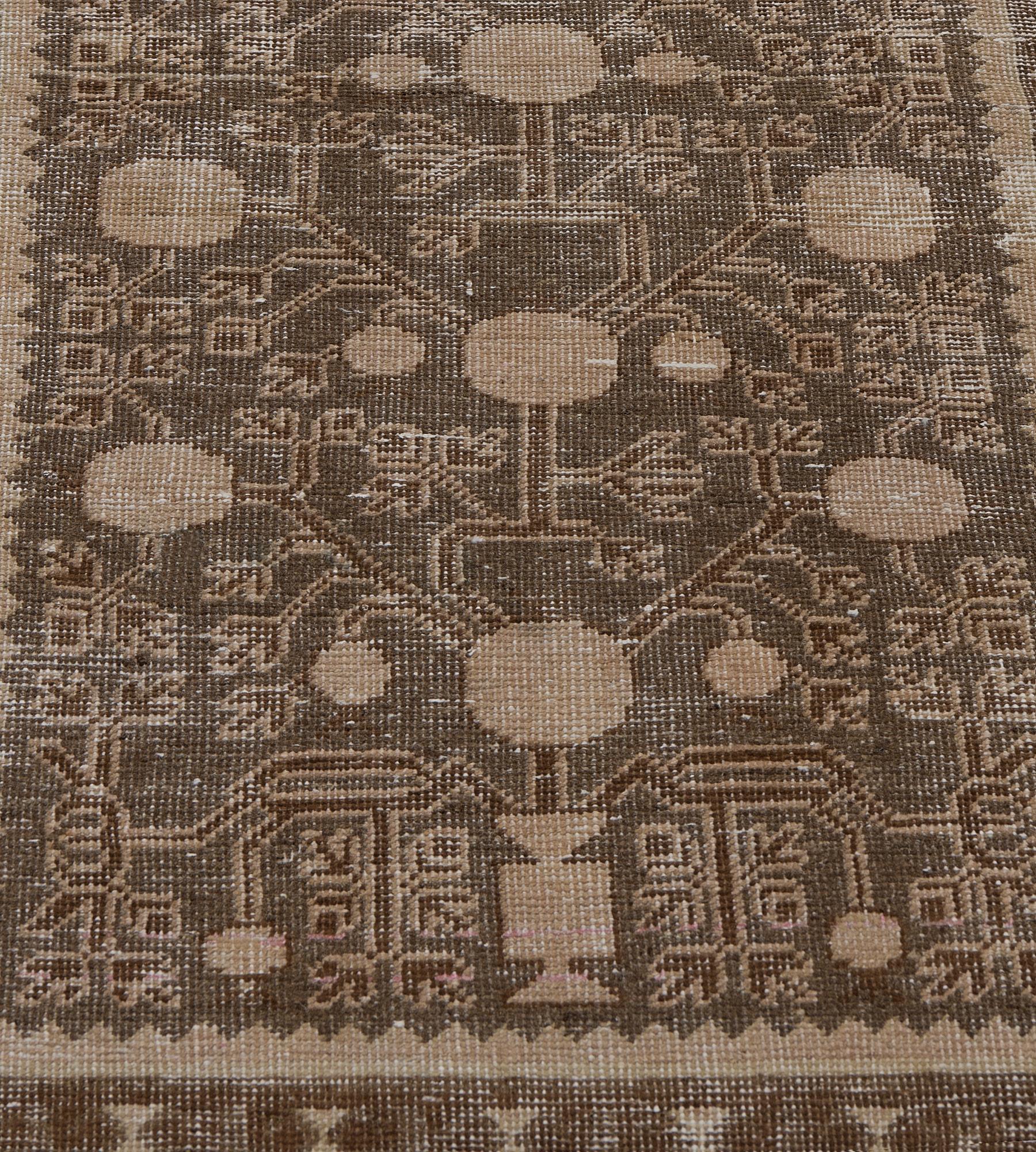 Questo antico runner di Khotan ha un campo marrone talpa con un vaso marrone bufala a ciascuna estremità che emette una vite angolare con melograni e spruzzi floreali, una striscia interna marrone bufala a zig-zag, un bordo a pannelli uncinati