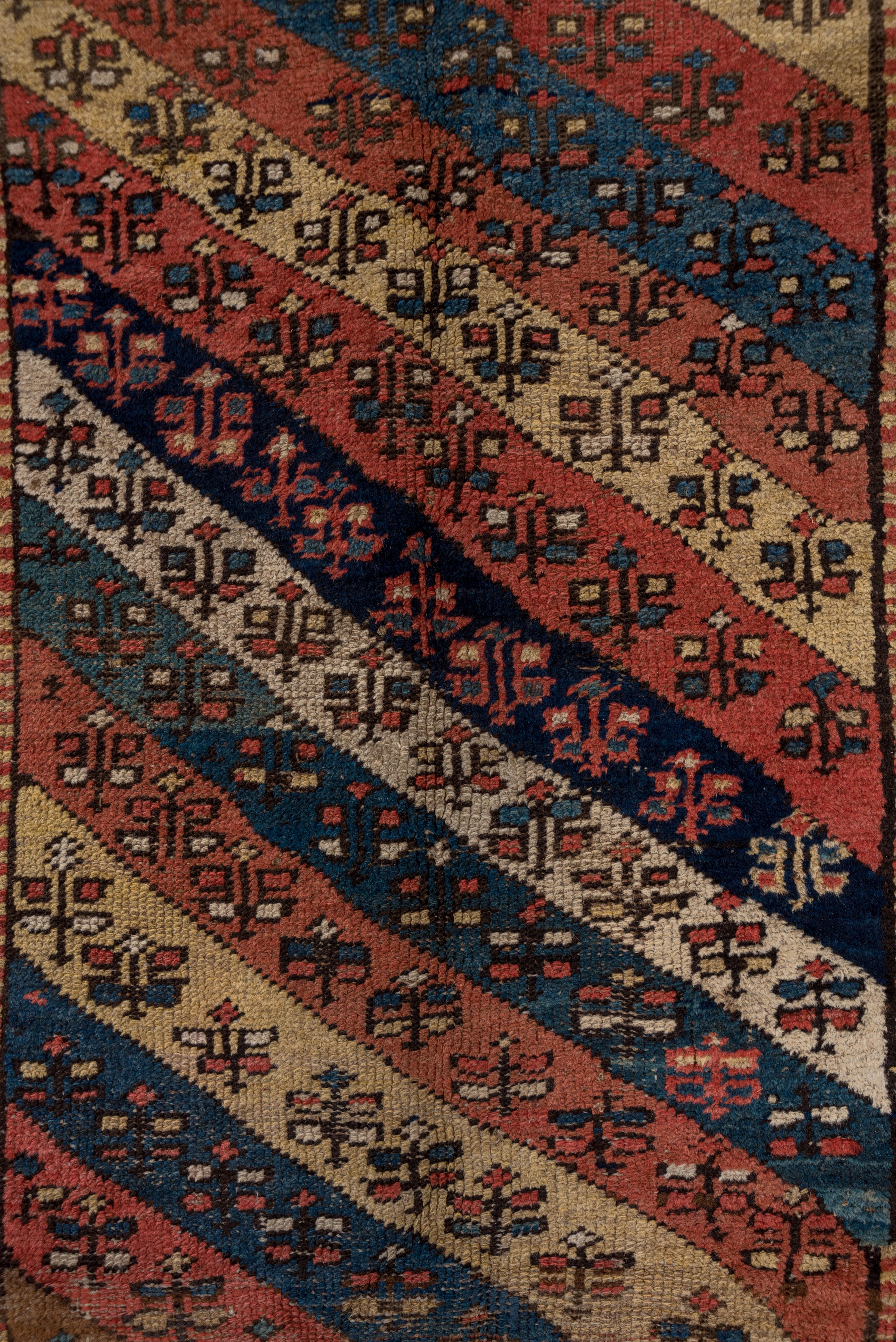 This fully geometric NW Persian village long rug features a diagonally striped field pattern in straw, ecru, navy, medium blue and coral, all with a small flower decoration. The outer border continues the slanted stripe idea while the main stripe of