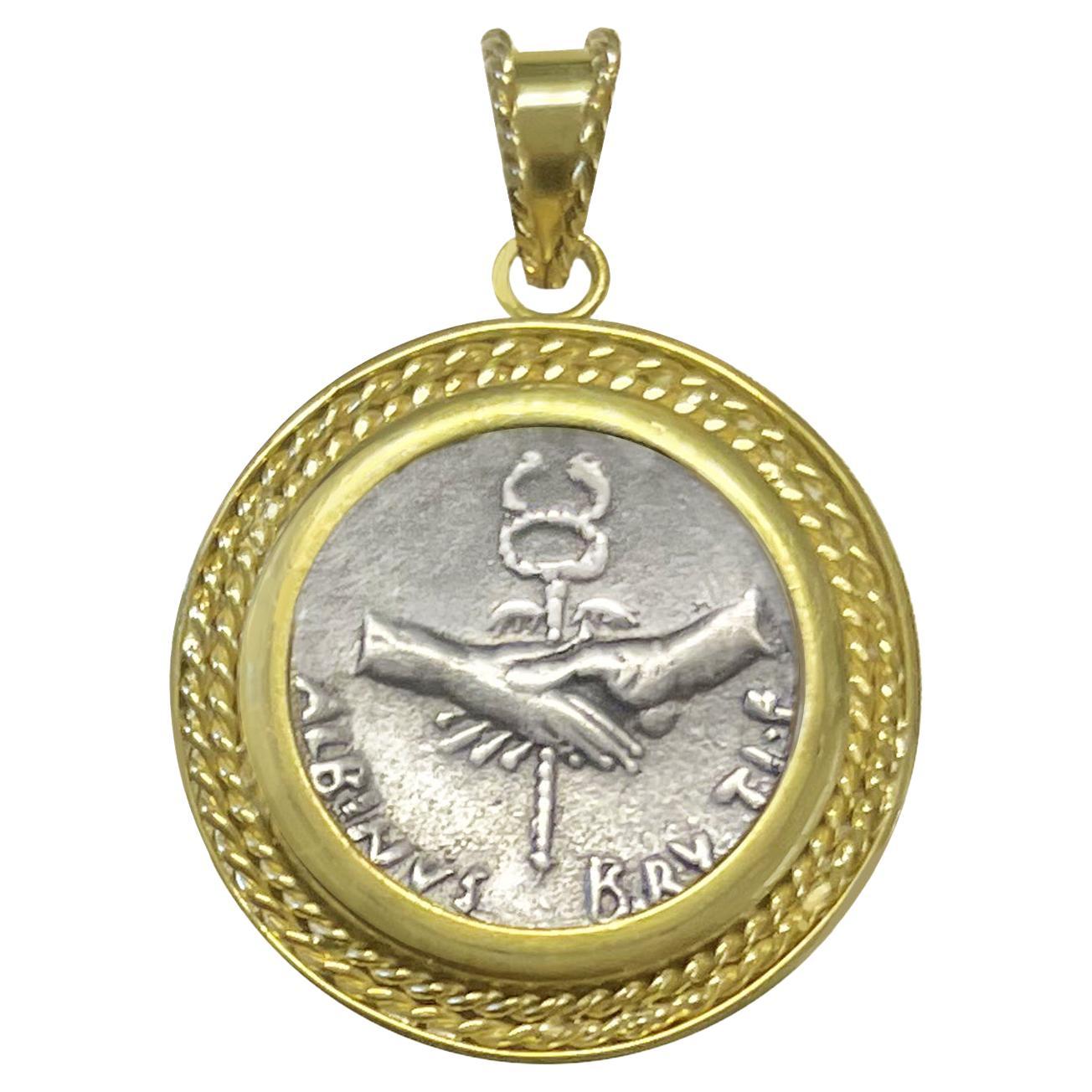 In this 18 Kt Gold pendant is set an authentic roman coin , minted by Albinus Bruti  ( 48 BC ) , depicting a Dextrarum iunctio: clasped hands holding winged caduceus. 
The hand shaking gesture, Dexiosis for the Greeks or Dextrarum Iunctio for the