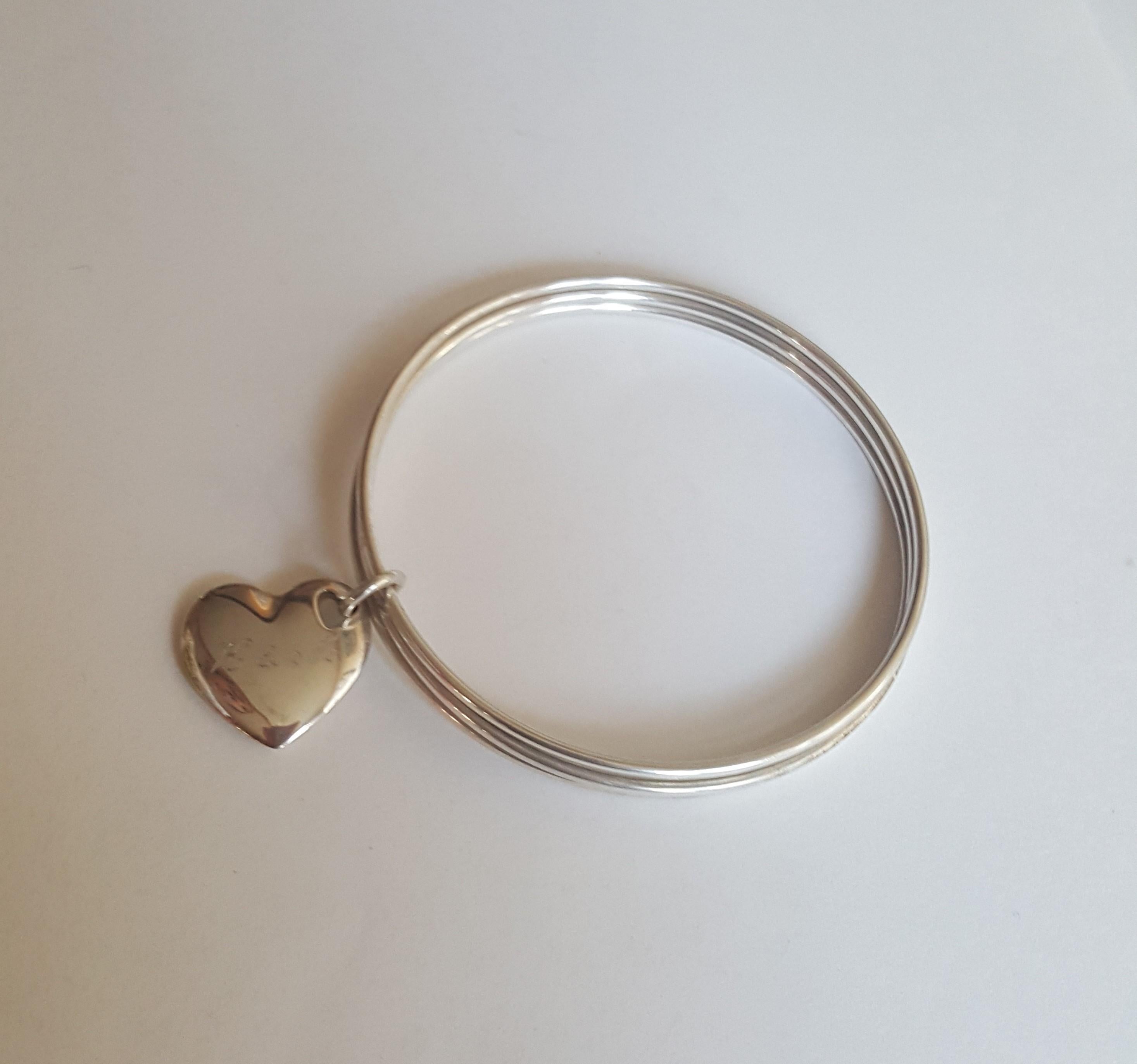 Authentic Tiffany Co Silver Trio Bangles with Tilted Heart Charm. Retired item. Very good condition.
This piece consists of a trio of slim silver bangles, and features a Tilted chunky heart charm. 24.2 grams. Standard Bangle Size. Each bangle is 2mm