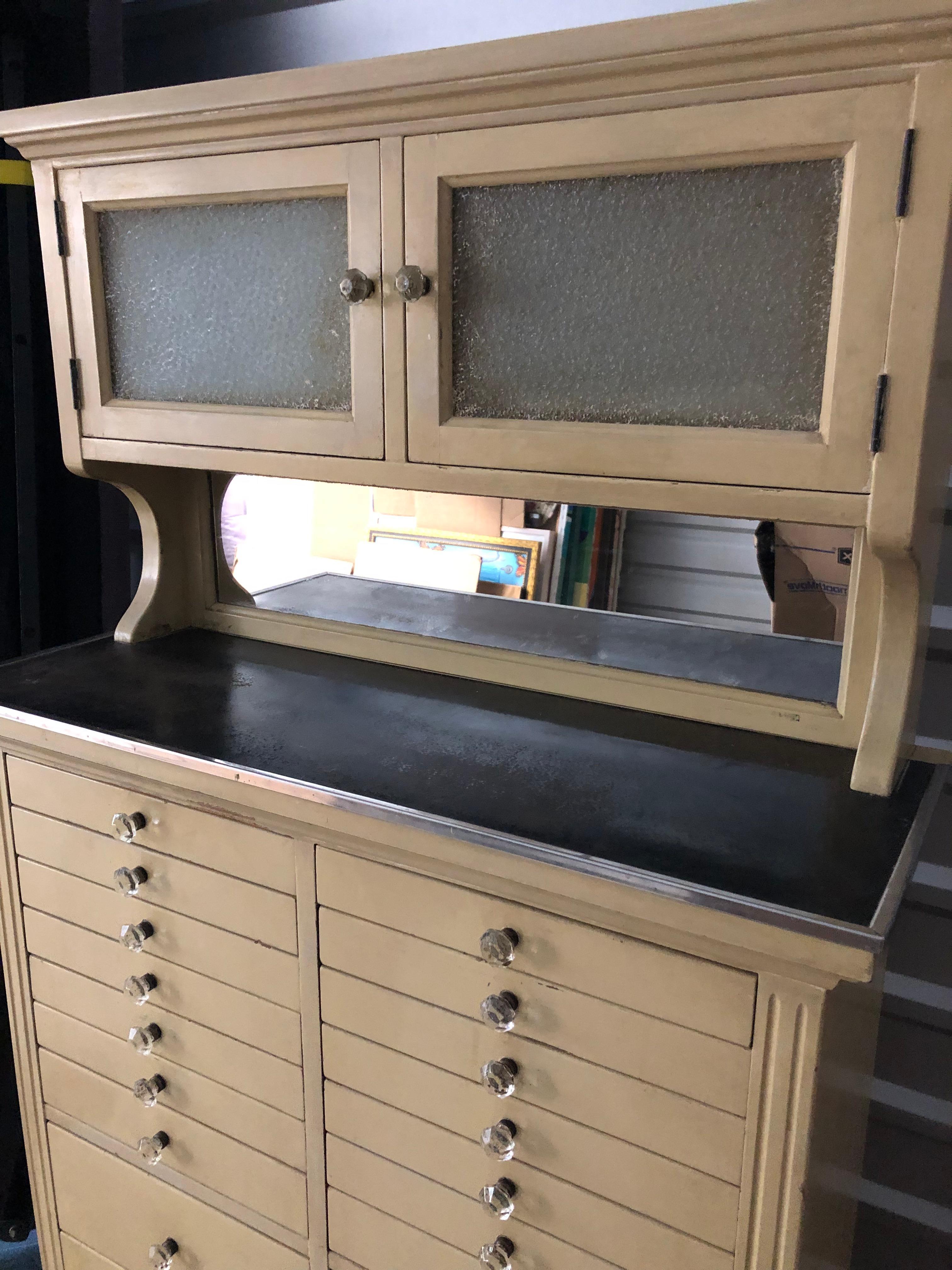 Incredible vintage American Cabinet Co. vintage dental cabinet having a multitude of drawers, two with original milk glass inserts.  The top area has a mirrored backsplash and two doors with opaque glass having storage inside.  The cut glass pulls