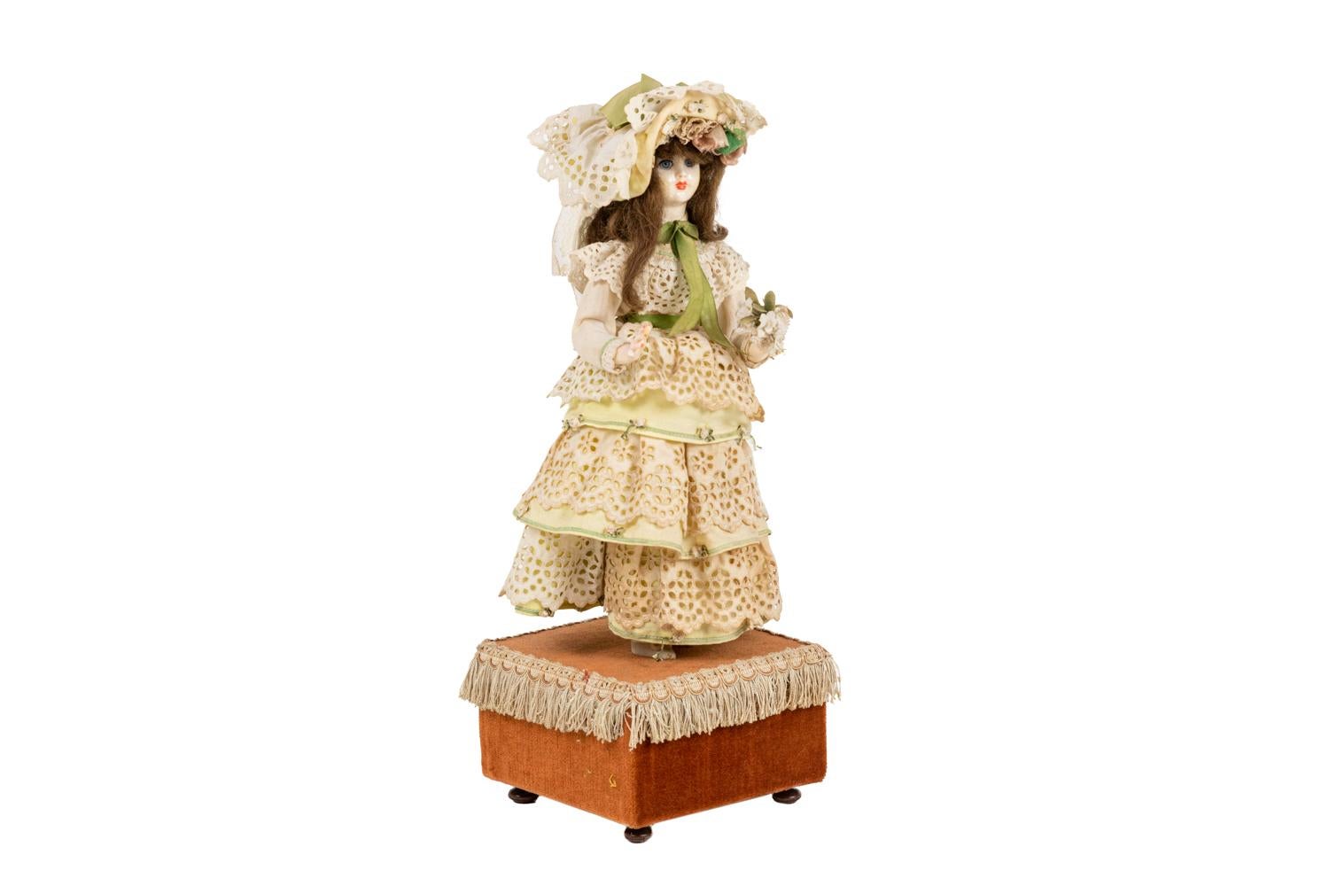 Automated doll, wearing a pale green dress with white lace and a hat in similar shades. She holds a small white flowers bouquet. A music box mechanism is hidden in the sienna-colored velvet base adorned with trimmings.

Work realized circa 1900.
