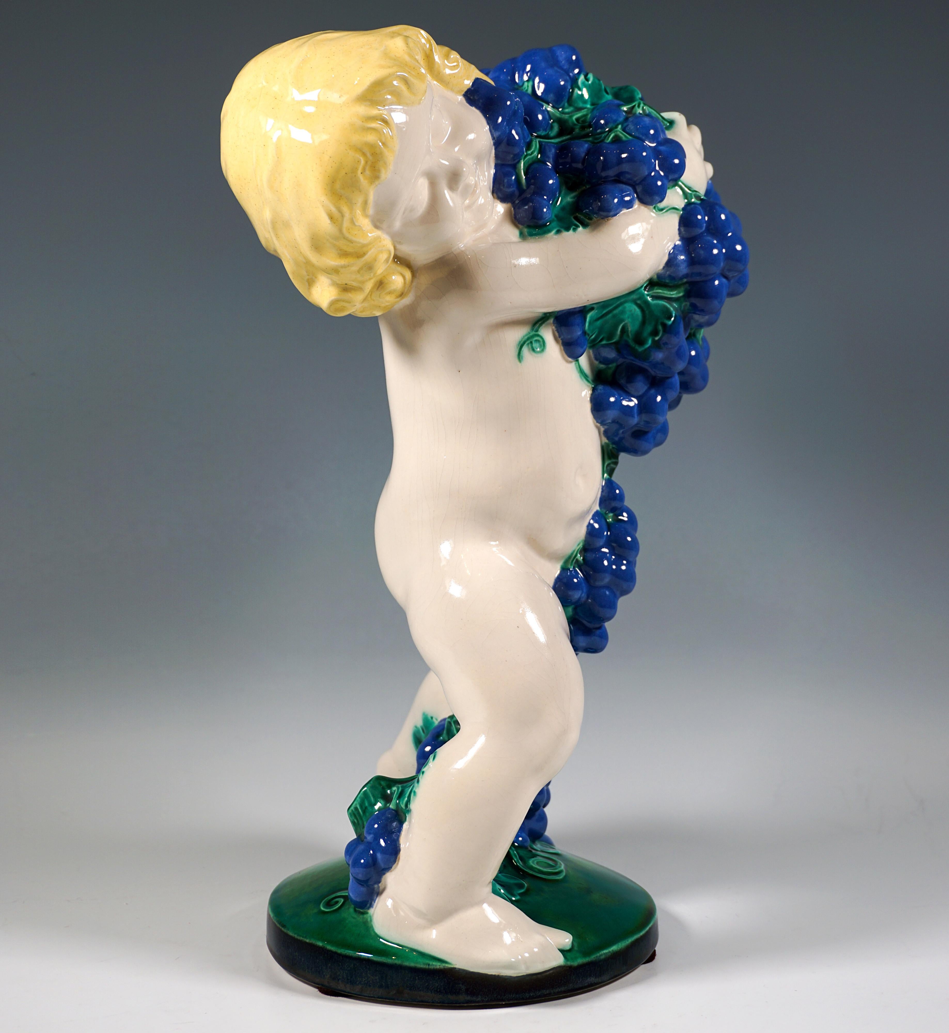 Excelente y rara pieza de cerámica Art Nouveau de una serie de las cuatro estaciones: 
Putto desnudo en posición de amplias zancadas, con la cabeza rubia inclinada hacia la derecha y sosteniendo una cascada de uvas que caen al suelo entre sus
