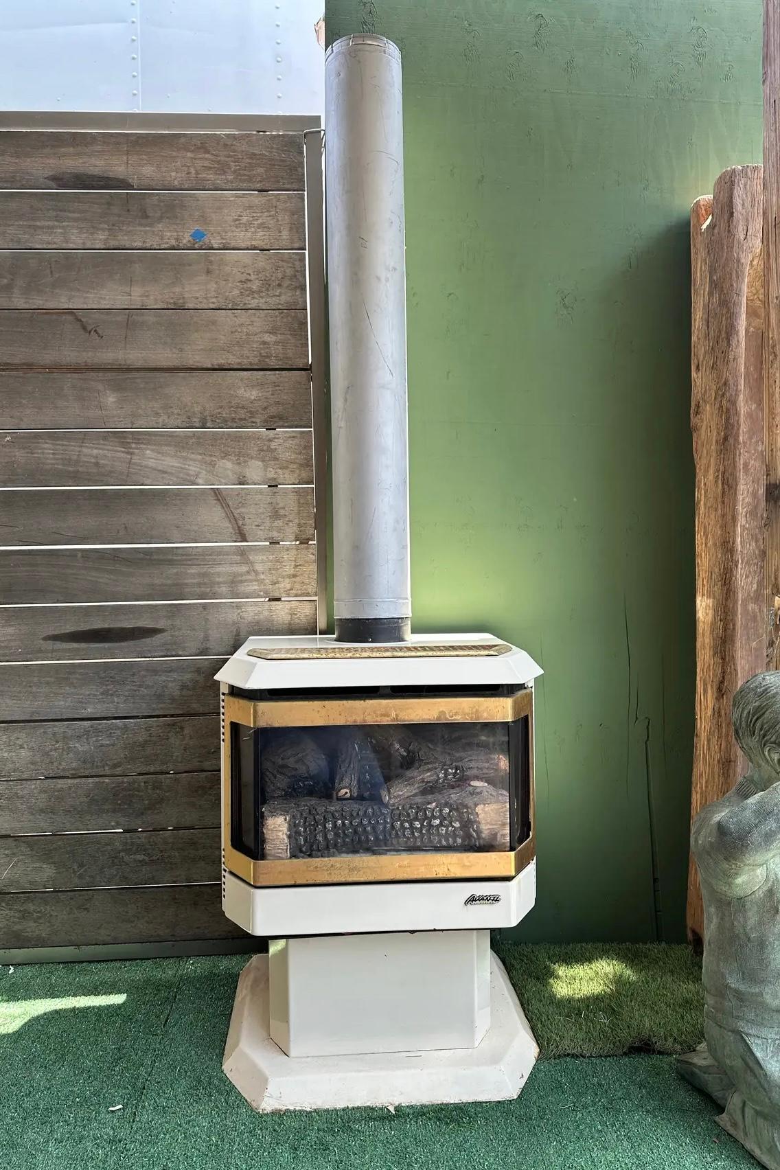Wonderful freestanding gas fireplace. It features a white base and top, with a glass front showcasing and artificial logs. It comes with three silver pipes extends from the top for venting, ( they are 36in H” each and all stick together are 108in