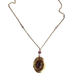 Avon 1970s Gold Tone Oval Locket Necklace with Purple Glass Accents (Collier avec médaillon ovale et accents en verre violet)