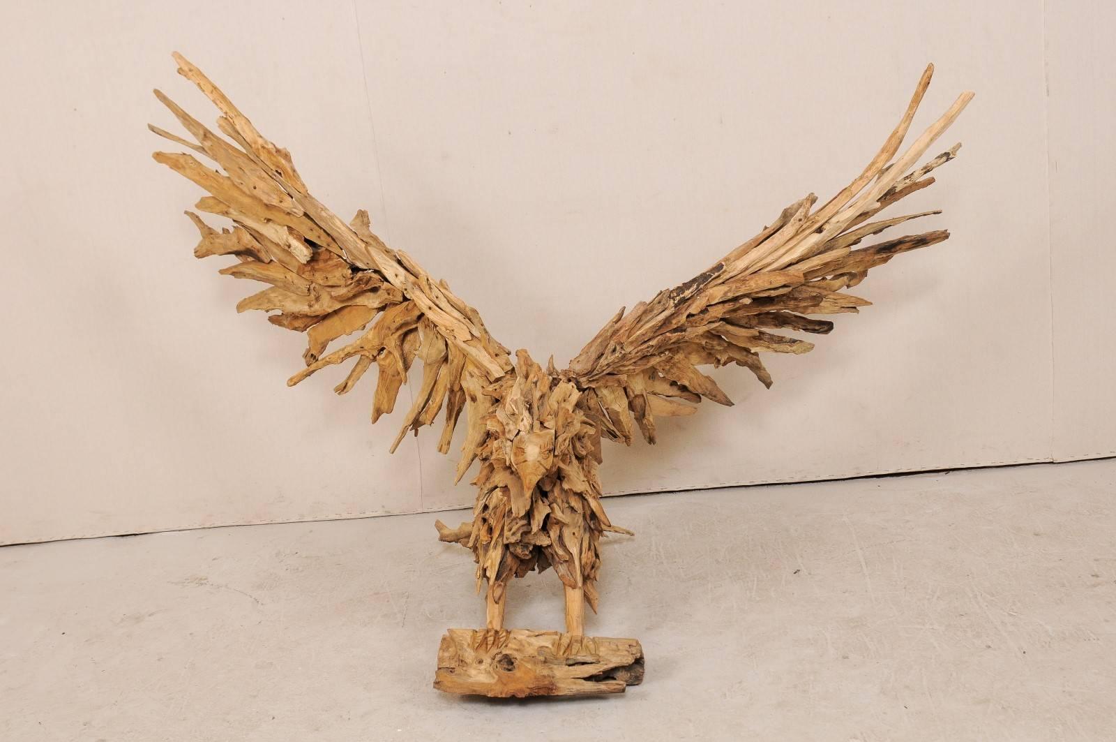 An awe-inspiring large-size eagle sculpture from driftwood. This majestic eagle is depicted with a broad upward wingspan, as if just landing or preparing for flight. The talons are gripped about a larger piece of wood, mouth closed, head straight.