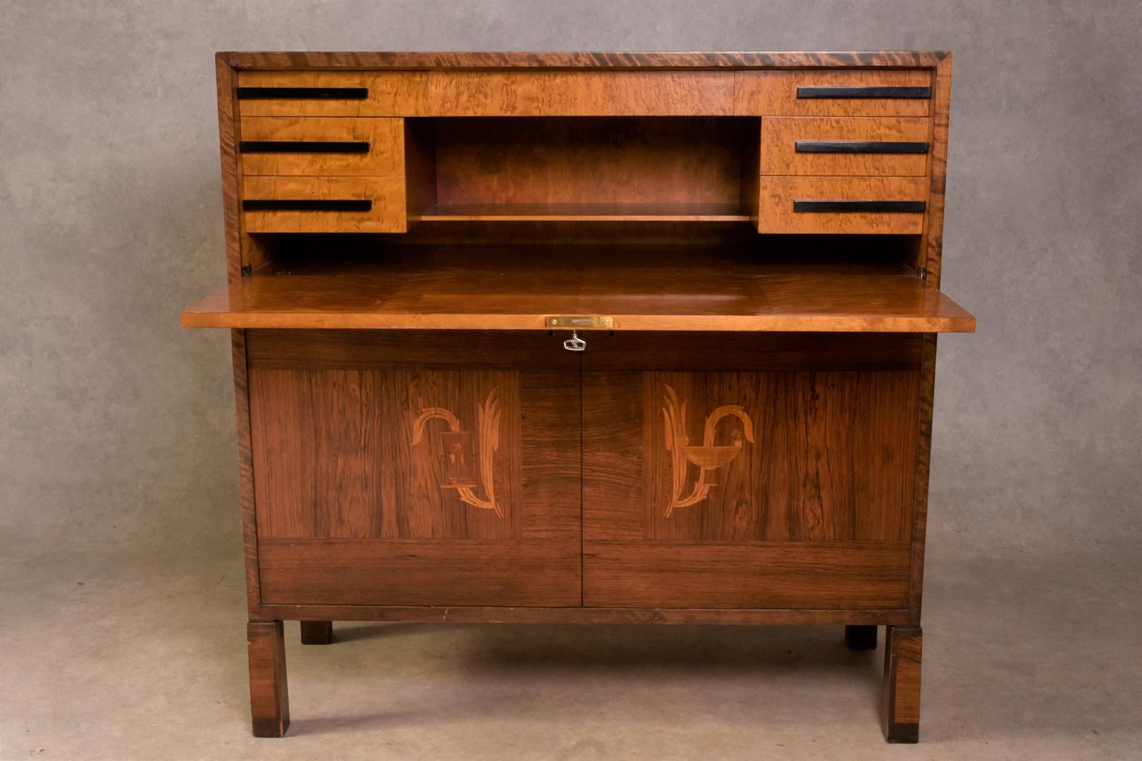 Cette armoire moderne suédoise a été conçue par Axel Larsson (1898-1975). Il est doté d'un rabat d'écriture et possède une serrure et une clé (clé incluse). L'armoire est en bois de bouleau et de chêne, avec un placage en orme. 
Un exemple