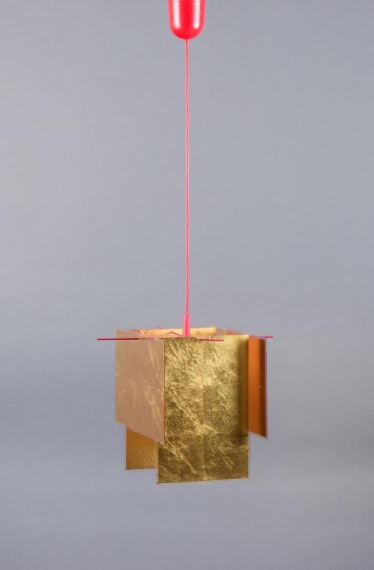 Axel Schmid per Ingo Maurer.
Pendente da soffitto con montatura in metallo laccato rosso.
Paralumi in plexiglass intarsiati con foglia d'oro a 24 carati.
Ciondolo modello 24 Karat Blau.
H. 26 cm. D. 28 cm. Corde lunghe circa 1 metro.
Fine del XX