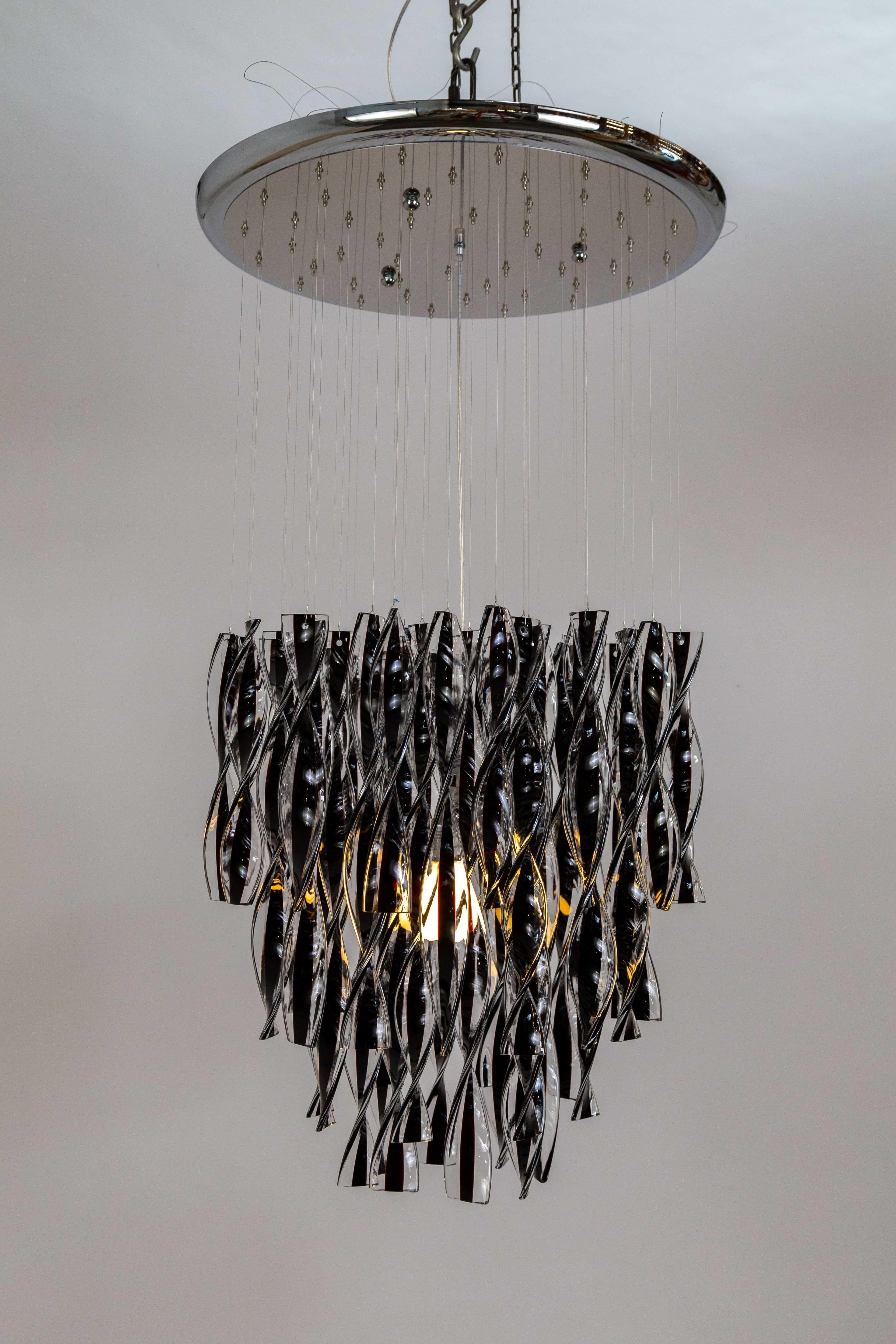 A waterfall pendant light of dangling black and clear glass ribbons reflect waves of light around it. A master glassmaker crafts the glass that composes a fluid and aquatic image with a diffused glow. The ribbons hang individually from a circular,