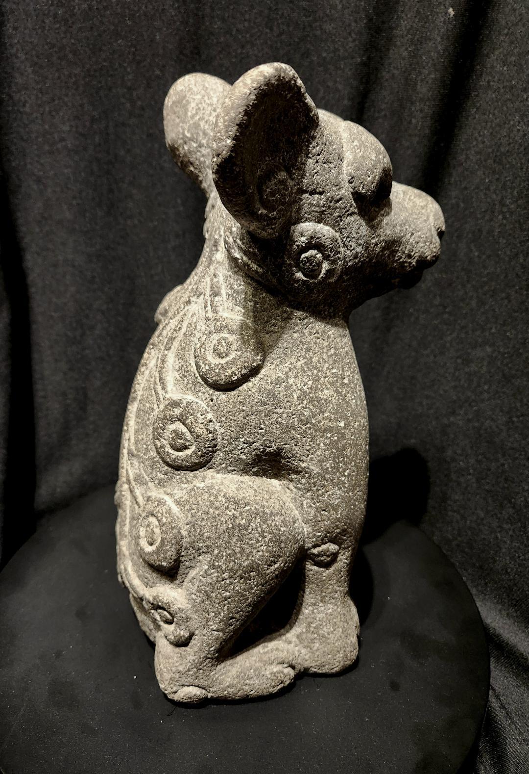An Aztec Stone Sculpture of a dog-like creature, seated with one raised paw, having large round ears, deep set eyes, mouth with exposed upper fangs, and an unusual curved tail with circular attachments.

This is an Aztec Water Dog, which is a