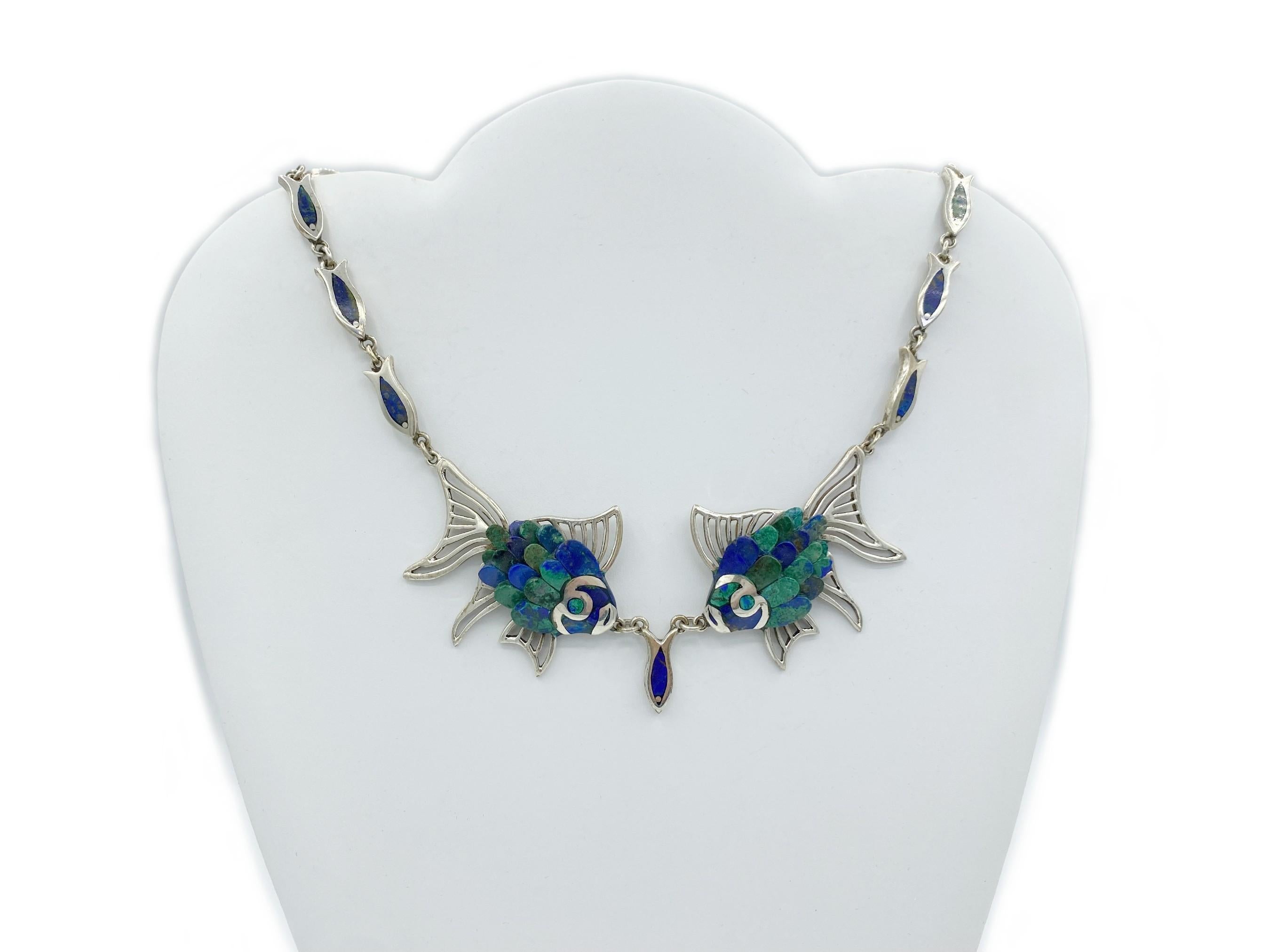 This unique, vintage Artisan necklace was handcrafted in Taxco, Mexico. Crafted of Sterling Silver, the eye-catching chain is comprised of linked fish-shaped panels of alternating inset blue azurite and green malachite, centered by two large fish