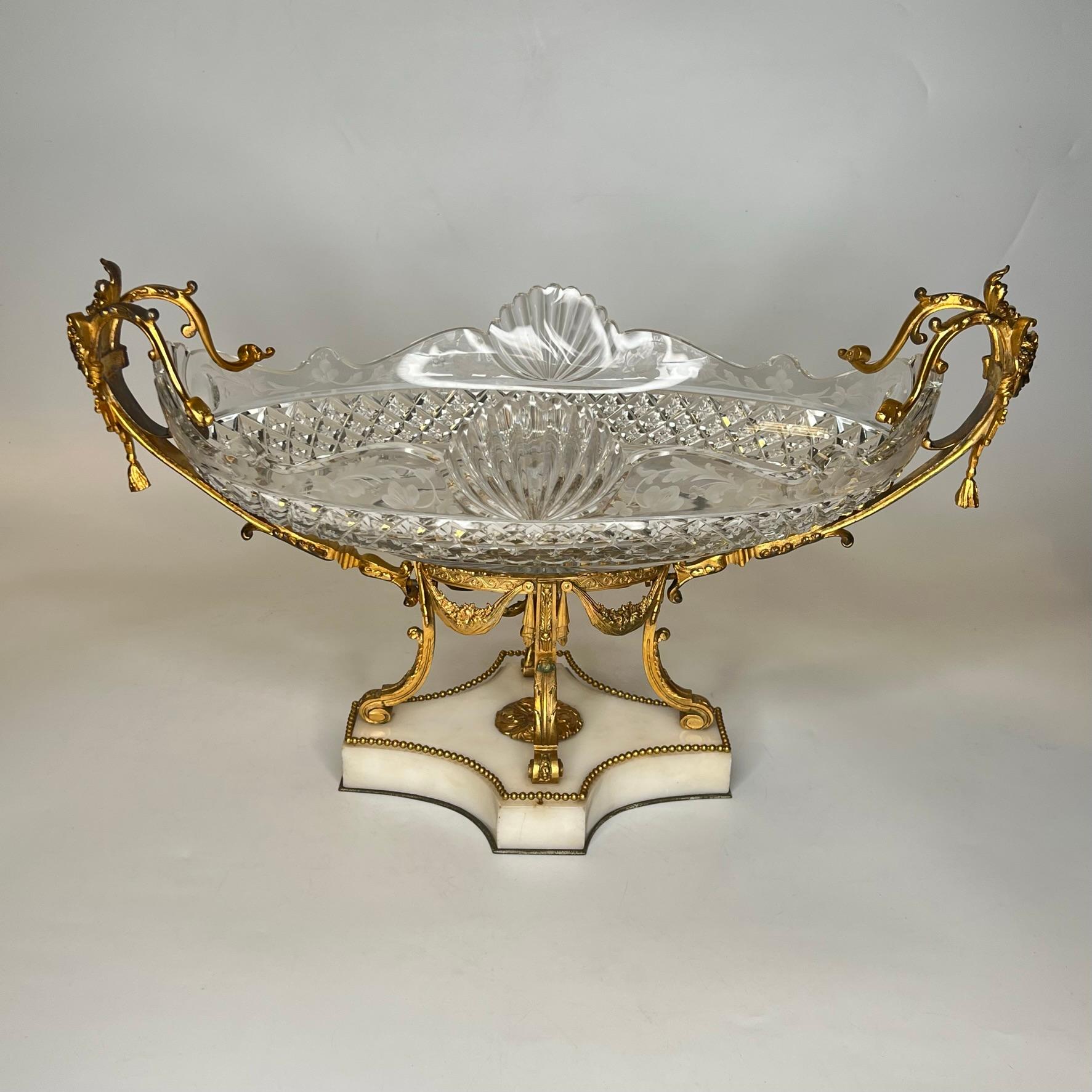 Notre centre de table français, vers 1880, comprend un grand bol en verre de forme ovale délicatement taillé avec des motifs de diamants et de coquillages et gravé de motifs floraux sur les côtés, ainsi qu'un cadre en bronze doré avec des masques de
