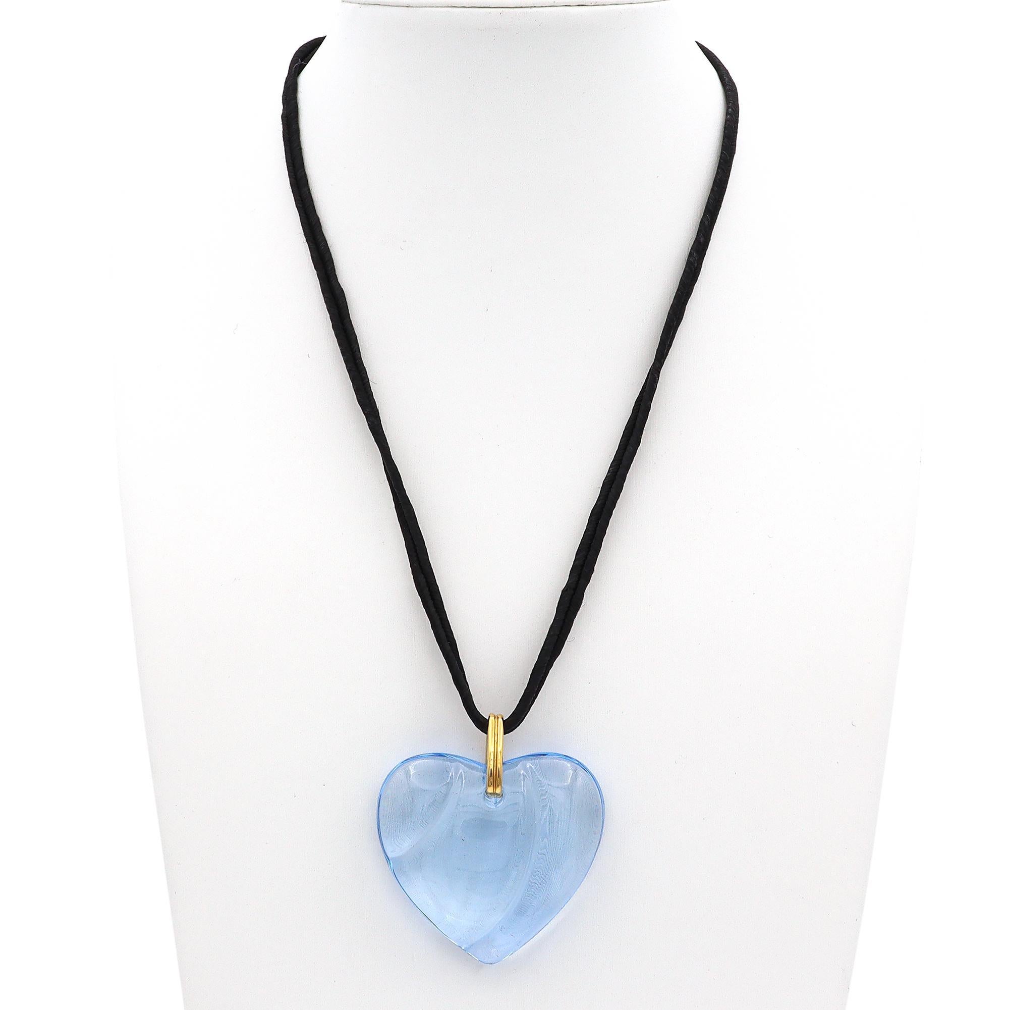 A stunning crystal pendant necklace by Baccarat. This exquisite piece features a heart-shaped blue crystal, showcasing a smooth and elegant design. The crystal is suspended from a simple black cord, accentuated by a gold-toned loop, adding a touch