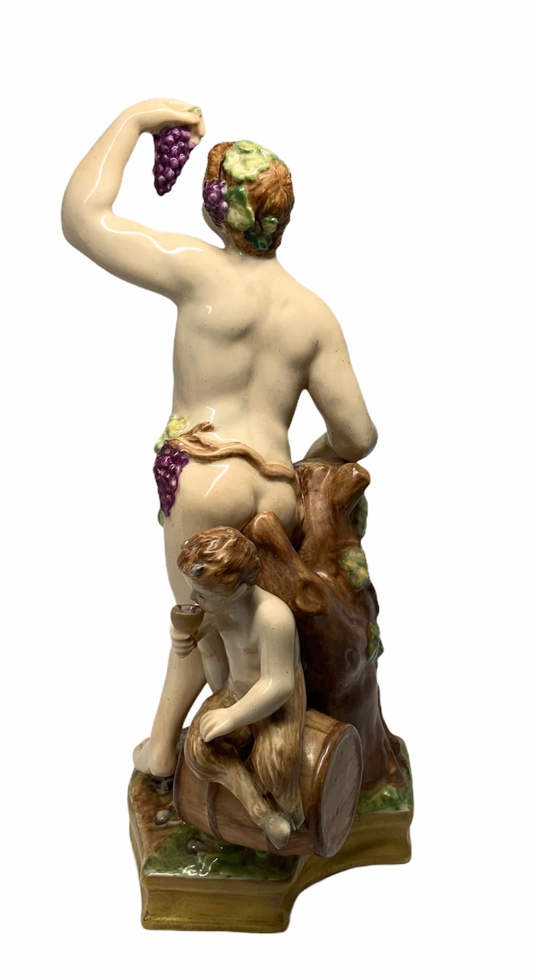 This is a hand painted porcelain of a semi nude Bacchus standing in front of a cut tree trunk while holding up a bunch of grapes. Behind him is a child faun drinking a cup of wine while sitting over a barrel of wine. There is a grape’s vine that