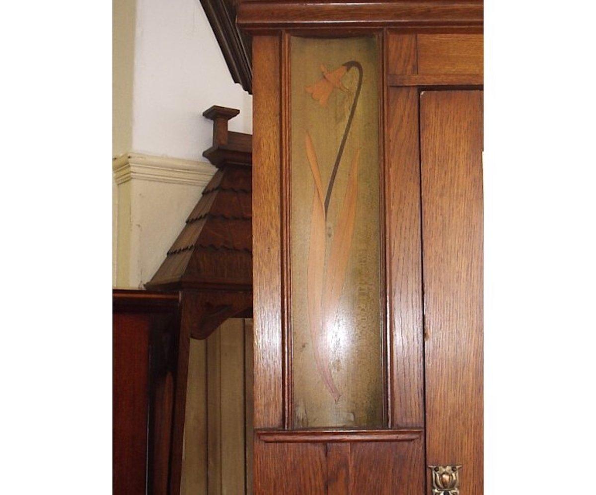 Shapland and Petter, in the style of Baillie Scott, attributed.
An Arts & Crafts oak armoire wardrobe, with a moulded flaring cornice and daffodil design inlays on concave panels to the upper sides and the original full-length bevelled mirror to the