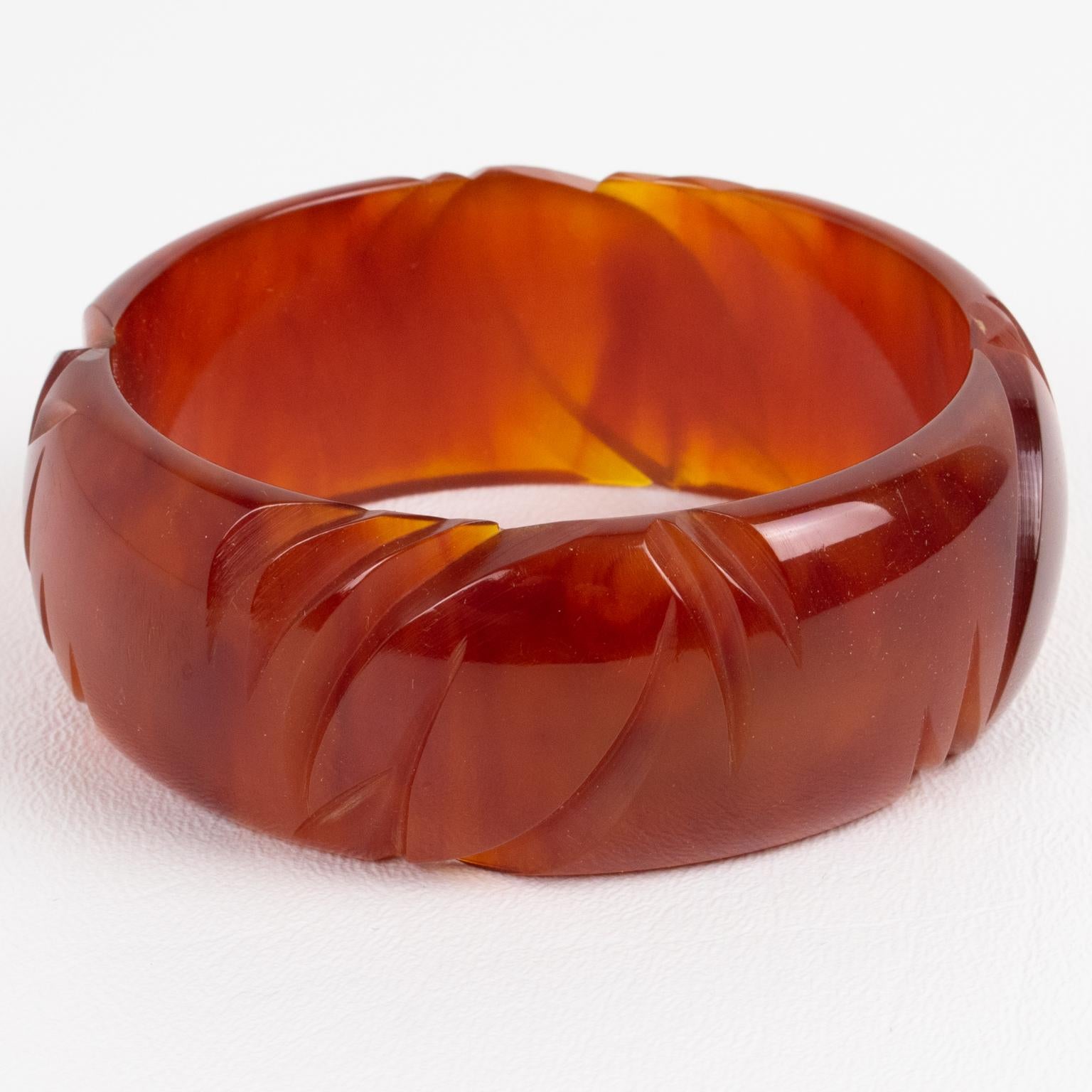 Se trata de una impresionante pulsera brazalete de mármol caramelo-ámbar tallada en baquelita. Presenta una forma abovedada con un profundo tallado geométrico alrededor. El color es un intenso tono caramelo marmóreo con remolinos y translucidez