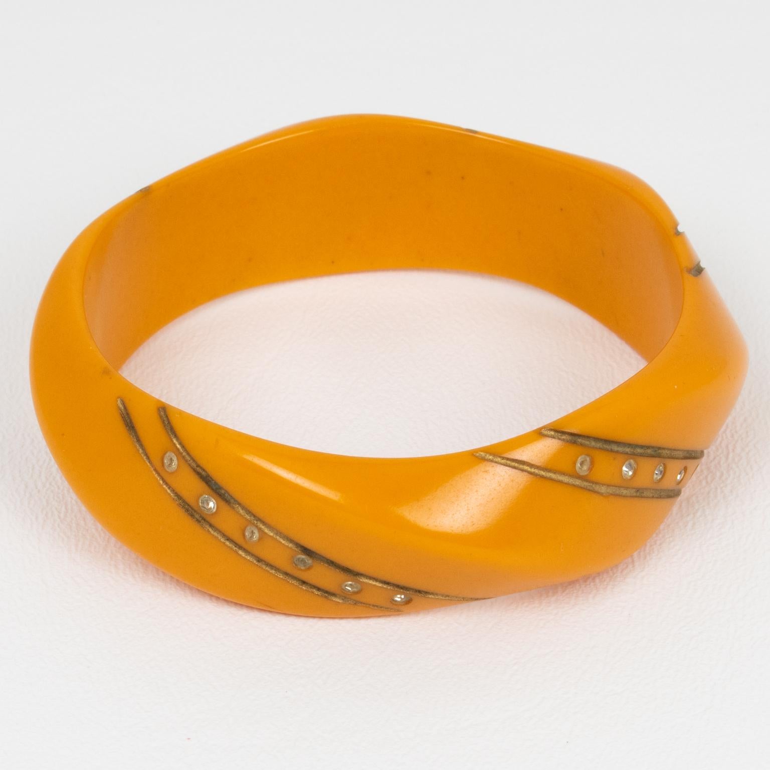 Questo raro e straordinario bracciale bangle in bachelite Art Deco presenta una forma a cupola con intagli geometrici a onda. È ornato da applicazioni di vernice dorata e piccoli strass di cristallo trasparente. Colore giallo/arancione intenso. 
Il