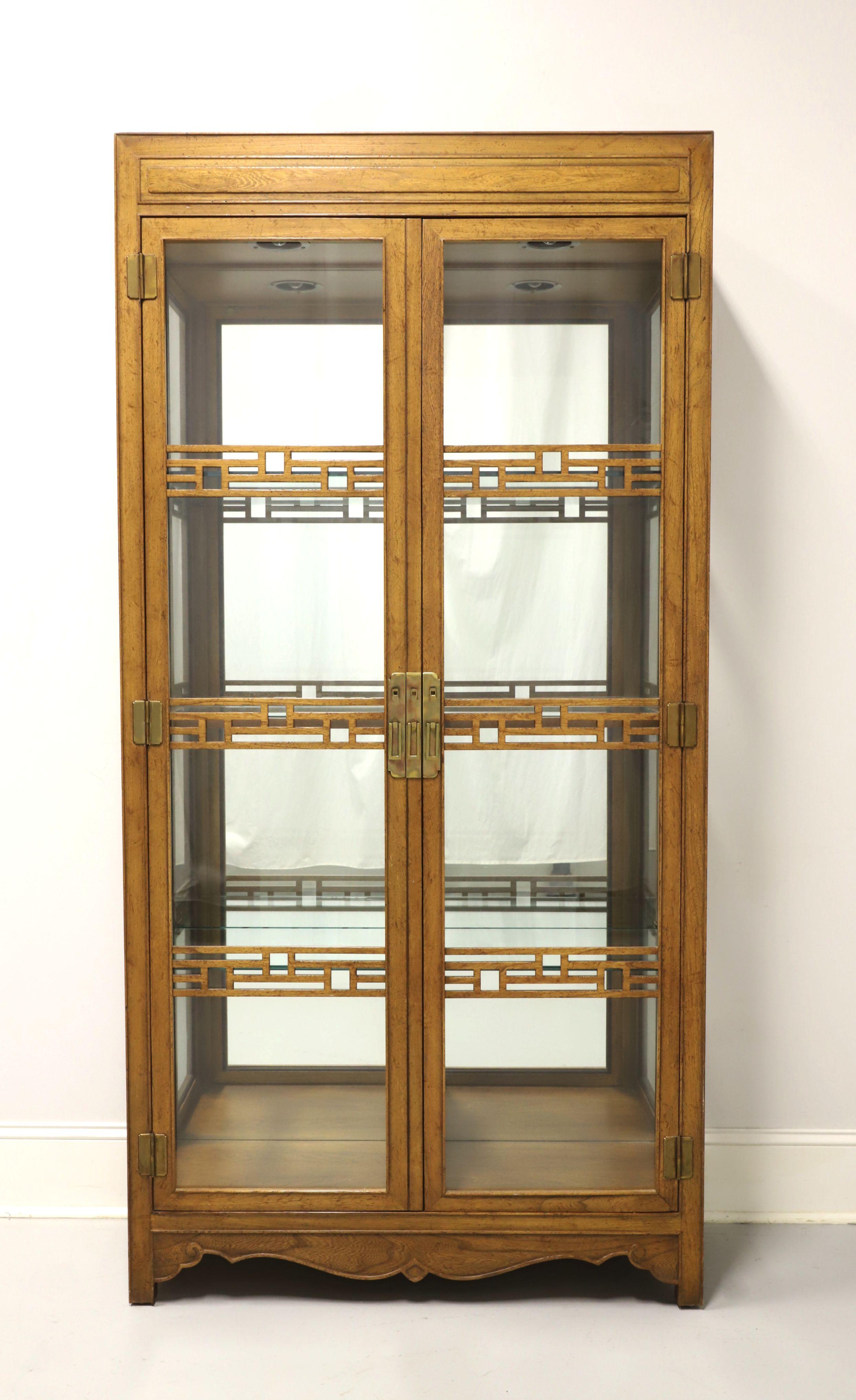 An Asian Style curio display cabinet by Baker Furniture. Oak with a light color distressed finish, brass hardware, Dual glass front doors with decorative chinoiserie fretwork and glass side panels. Features lighted interior cabinet with dimmer