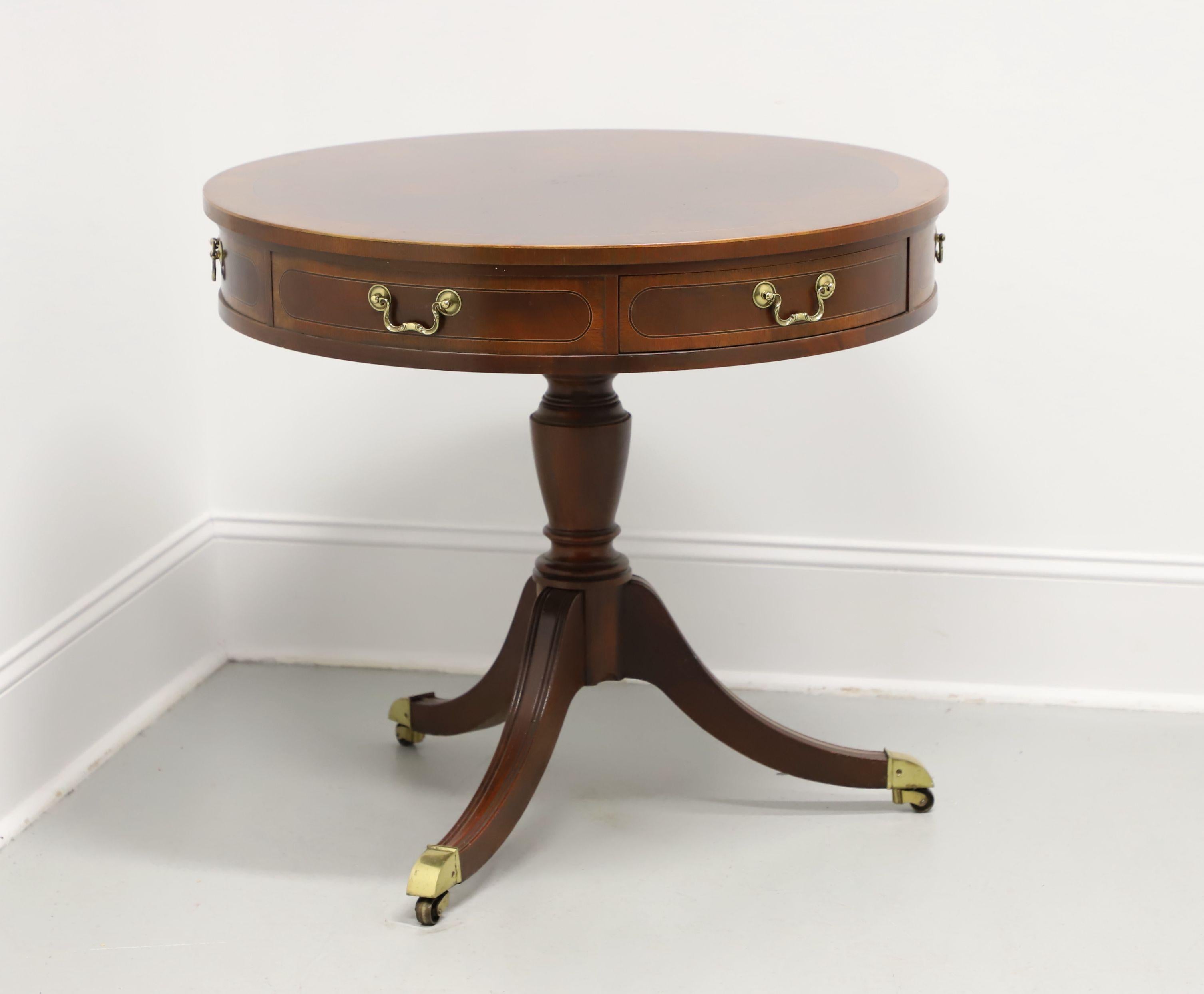 A Federal style round drum table by Baker Furniture. Mahogany with brass hardware, banded rounded edge top with inlaid flame mahogany, banded drawer fronts, turned pedestal base, three fluted legs with brass toe caps, and brass casters. Features two
