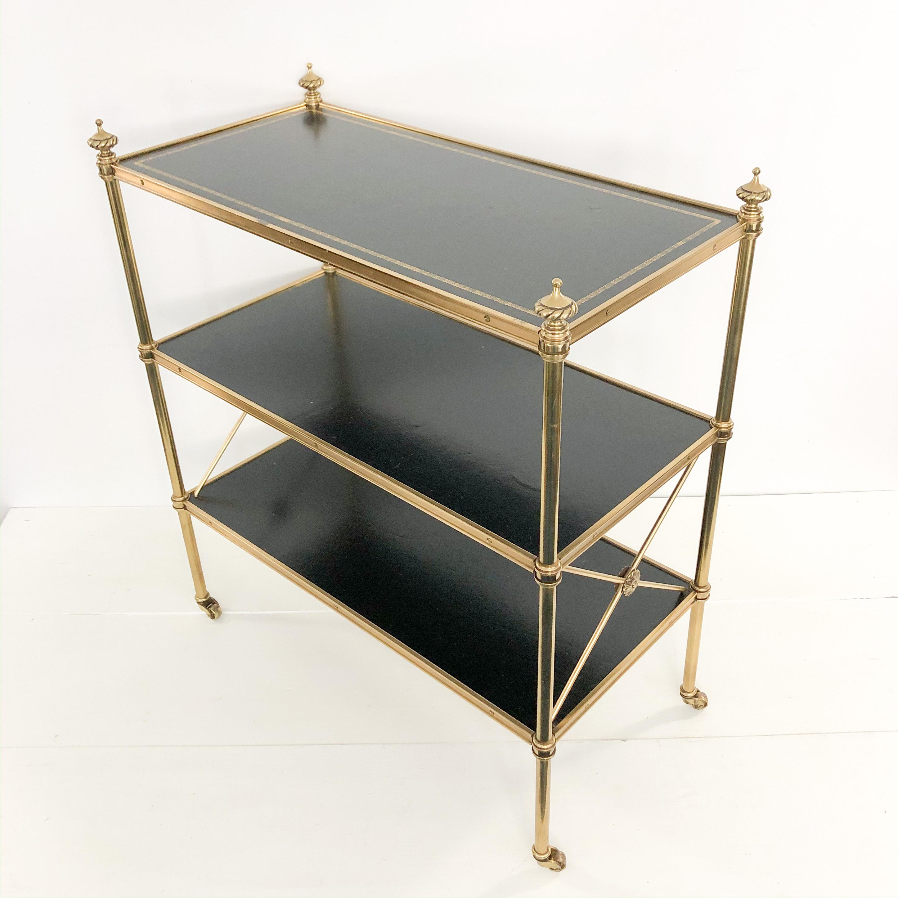 A Baker Directoire style solid brass three-tier rolling cart / shelving / bookcase / étagère with gilt embossed leather-topped shelf from the 1950s-1960s. It has the metal Baker Furniture Company metal label from that era on the underside of the