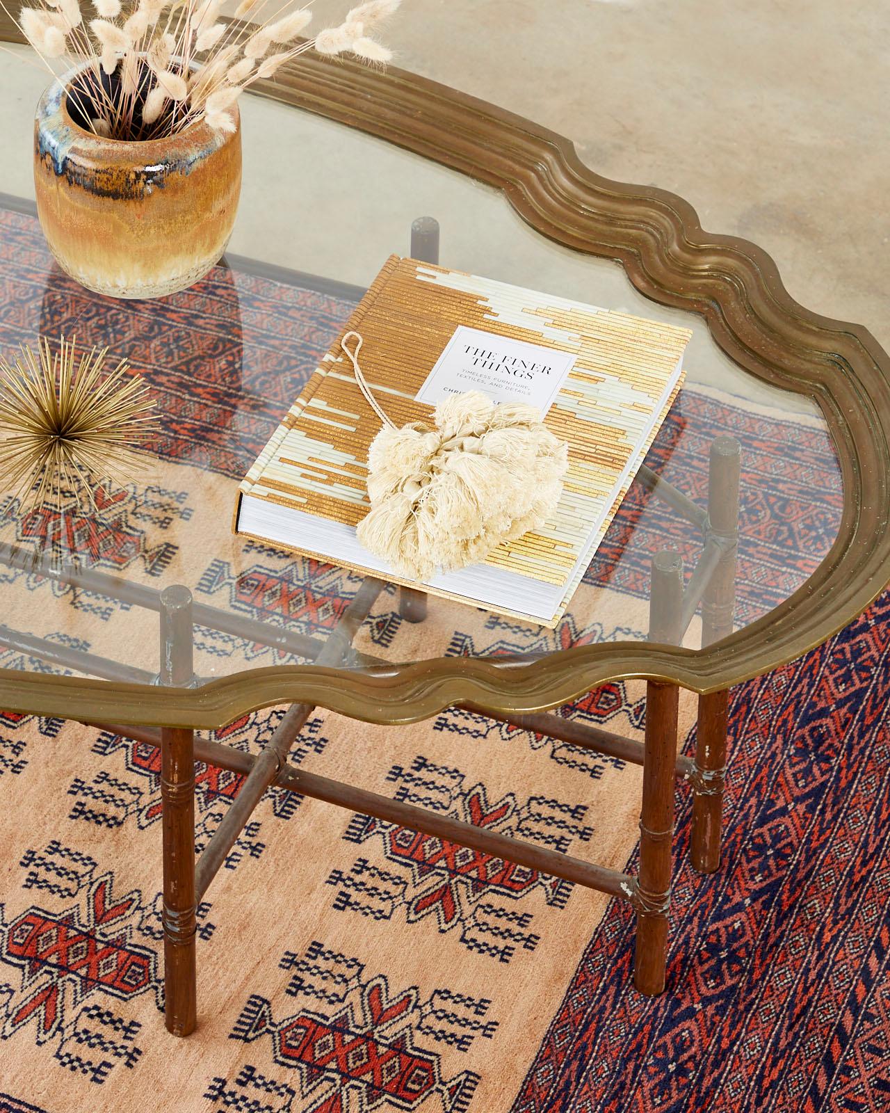 Hollywood Regency brass and glass tray top coffee or cocktail serving table. Features a faux bamboo metal base supporting a large brass tray table with scalloped edges framing a pane of glass. The mid-century modern tray was offered by Baker