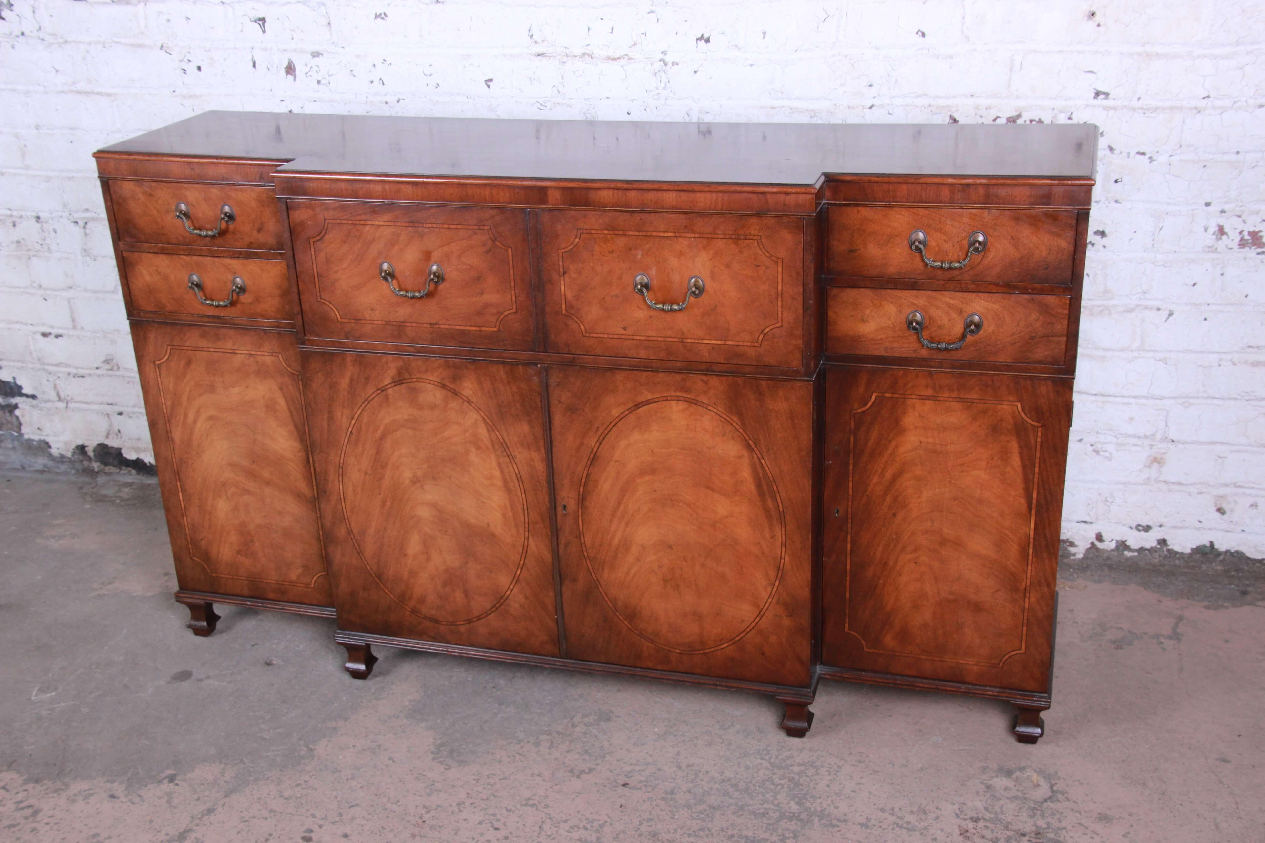Offering a nice Baker Furniture banded mahogany sideboard or credenza. The credenza has a nice brown mahogany finish with inlaid banded designs and the drawer and doors. The top features two drawers on each side with a large drawer for ample storage