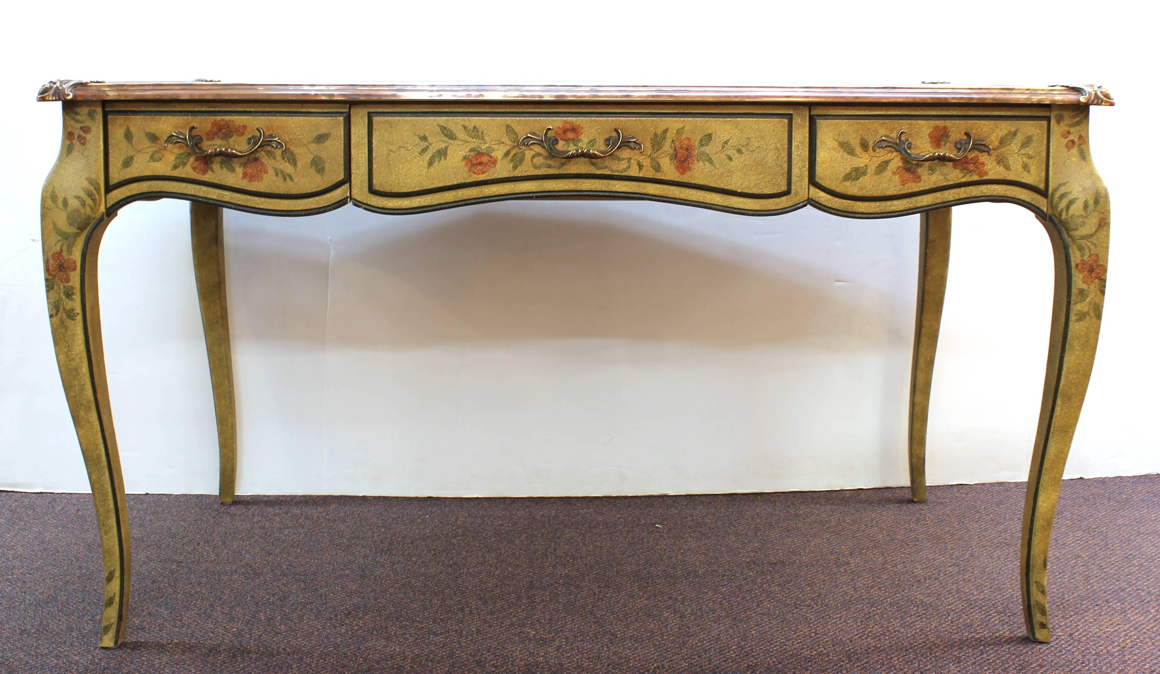 A baroque style writing desk made by Baker Furniture, with a decorative floral and foliage background and a leather top set inside a patinated brass frame. The front has three dovetailed drawers. In good vintage condition with age appropriate wear