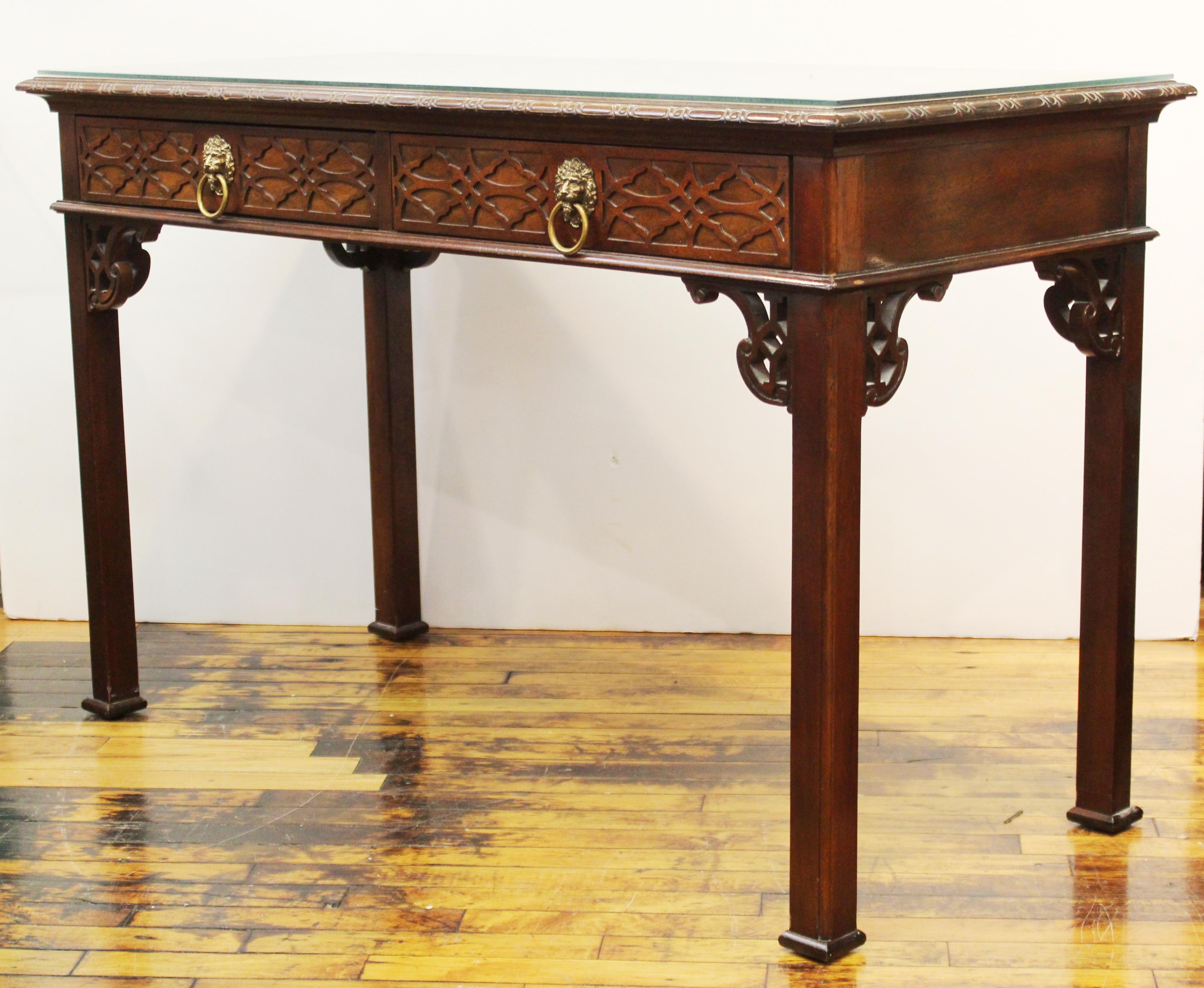 Midcentury Chinese Chippendale style writing desk or console table in mahogany finish with two drawers with lion's head pills and a removable glass top, designed by Baker Furniture Company. Metal oval plaque with makers mark located in one of the