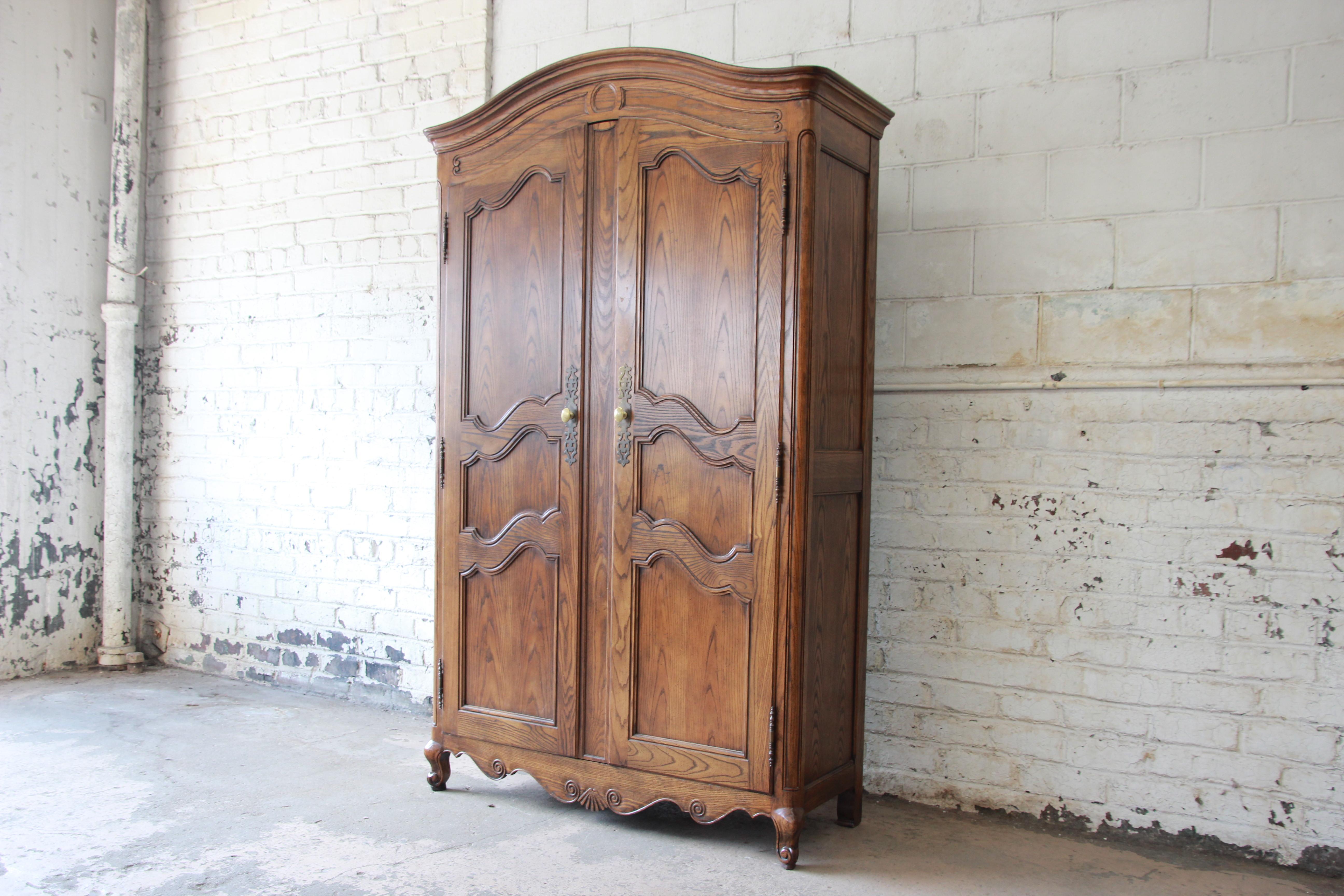 A gorgeous French Provincial Louis XV style armoire or wardrobe by Baker Furniture. The armoire features beautiful carved wood details and solid oak construction. It offers ample room for storage, with three open shelves, three cubbies, and three