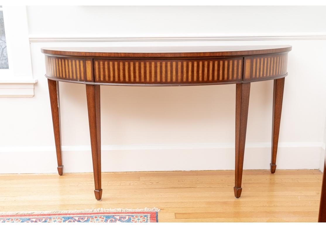 Baker Furniture demilune with shell design, pie cut figured mahogany with cross banding. The frieze with alternating marquetry inlay and resting on tapering legs with spear feet.

Dimensions: 19