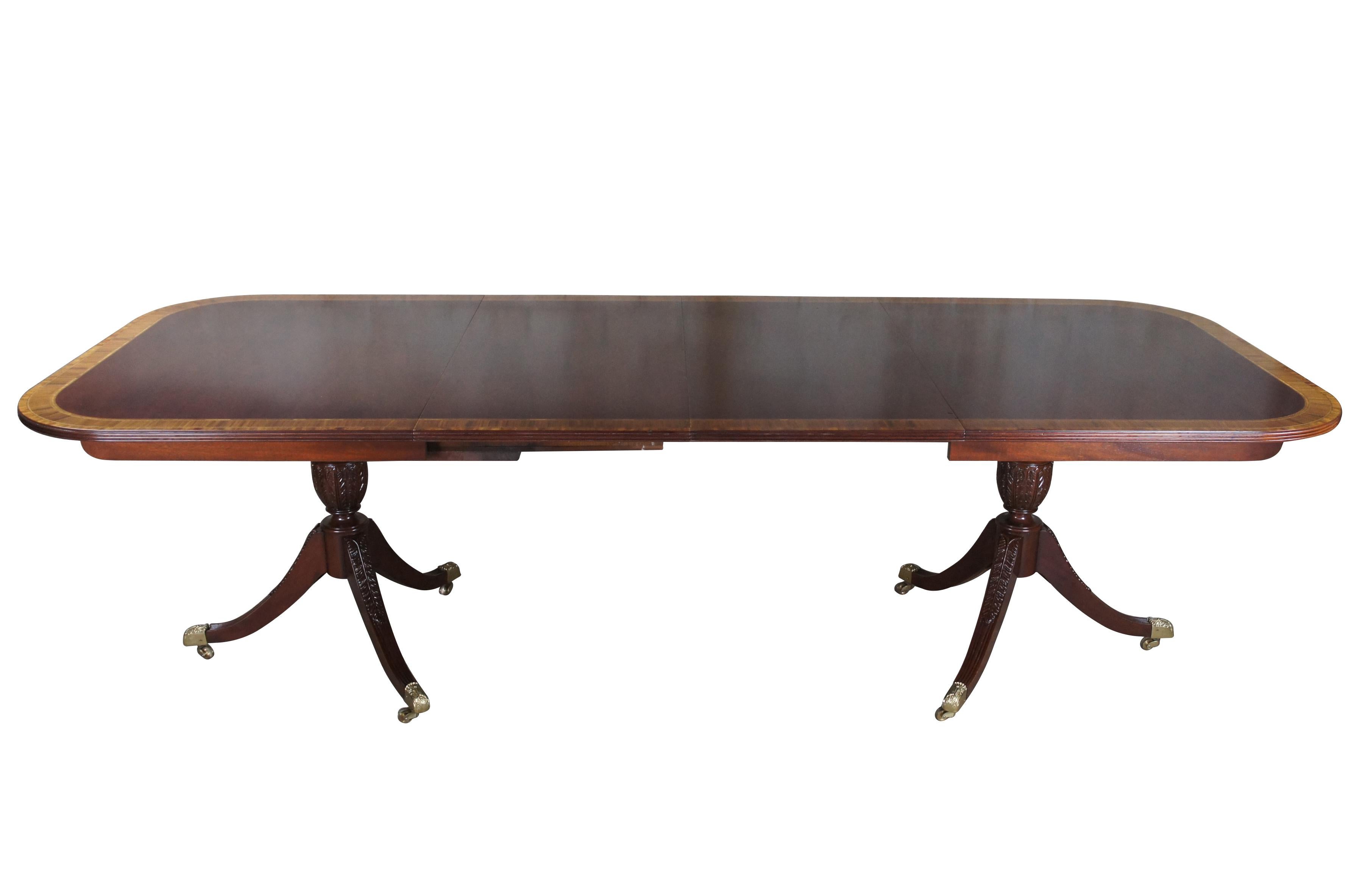Baker Furniture English Regency style dining table. Features an impressive mahogany banded top with two leaves for extension and table pads. The table is supported by a double pedestal mahogany tripod base with shapely acanthus carved urns over