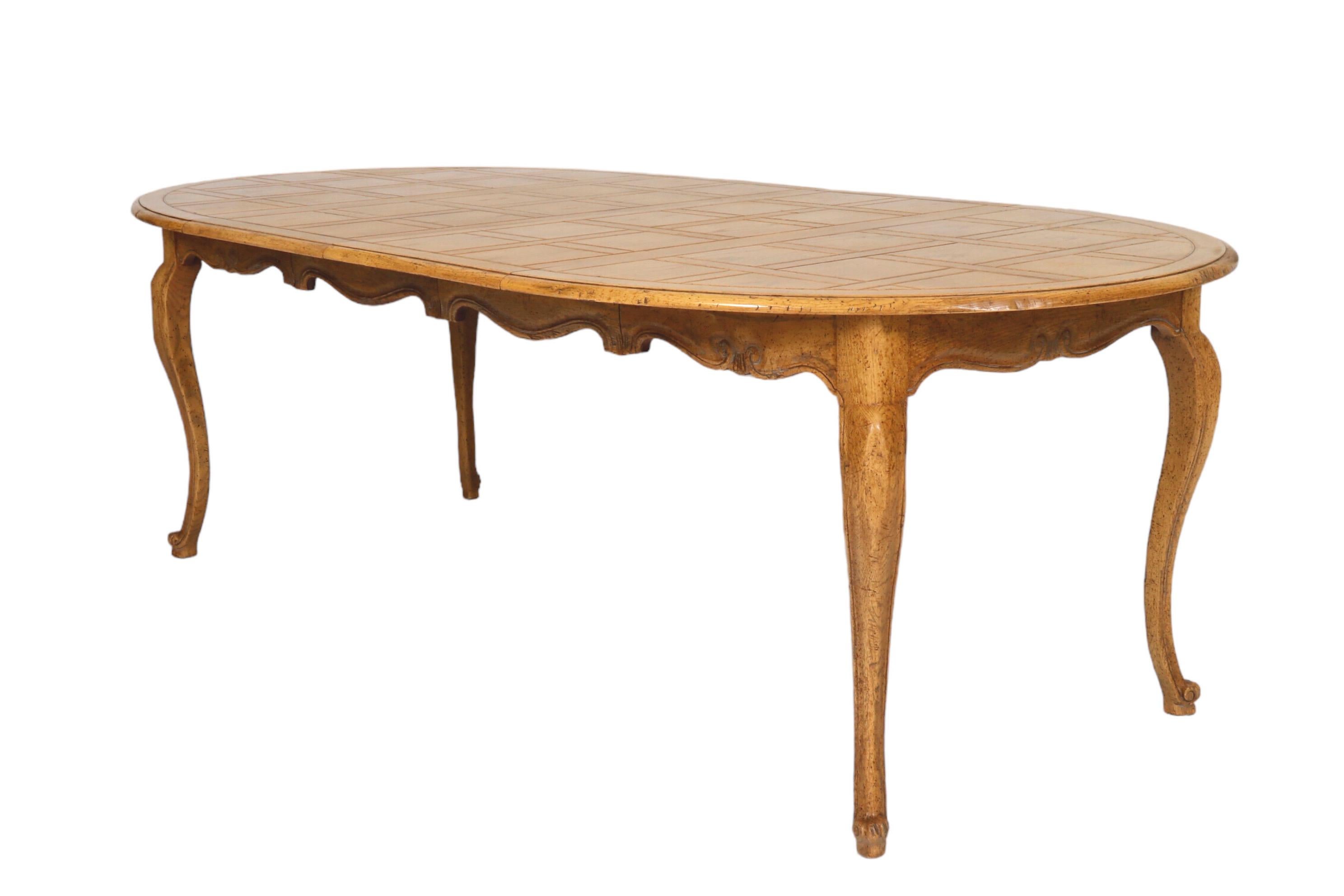 An oval French Country dining table made by Baker Furniture. The tabletop is decorated with carved lines in a Chantilly pattern. Below, a serpentine table skirt carved with s-scrolls along the edge meets four cabriole legs finished with whorl feet.