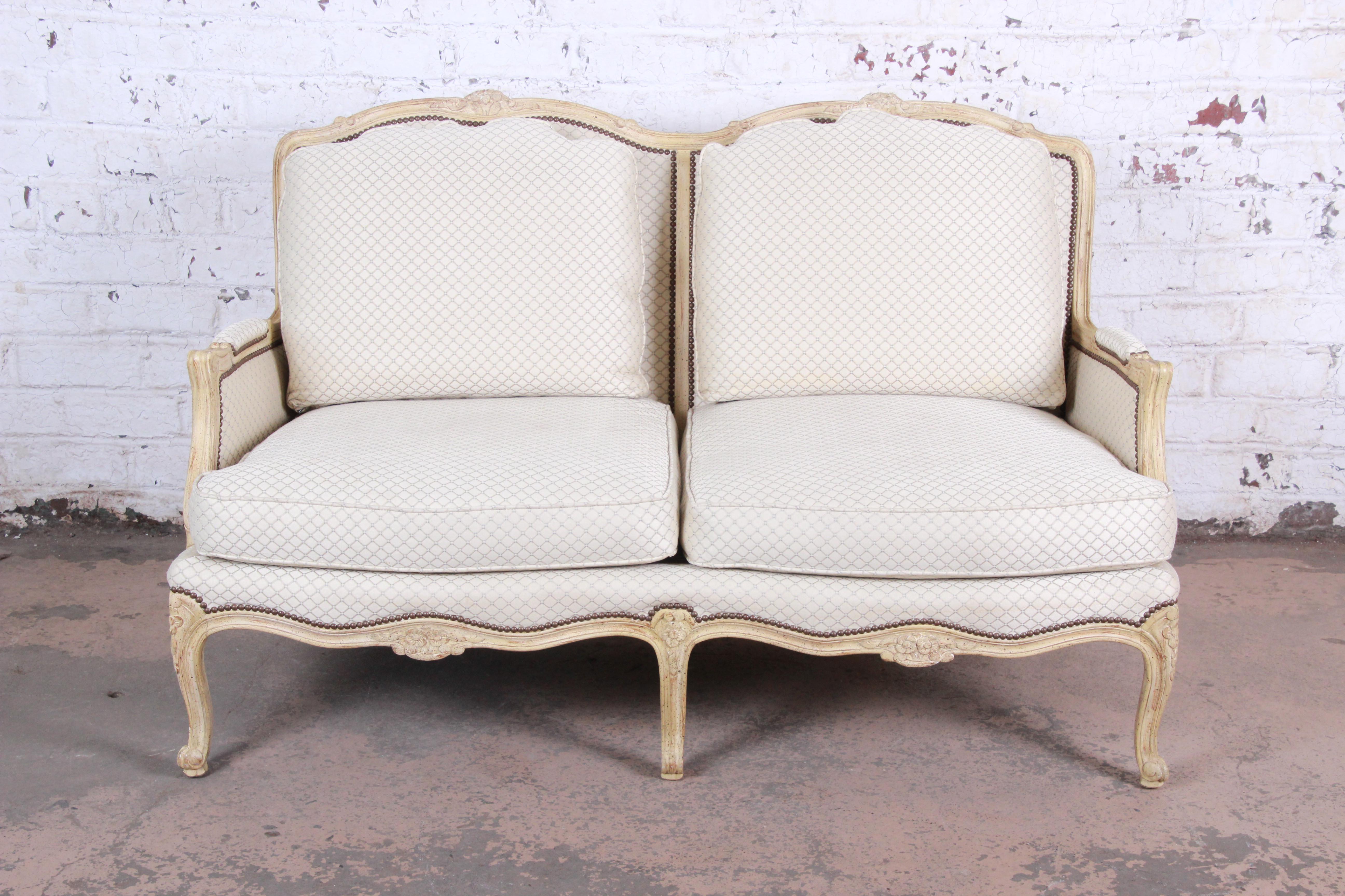 A gorgeous French Provincial Louis XV style loveseat or settee by Baker Furniture. The loveseat features stunning carved wood details with cabriole legs and studded ivory upholstery. The upholstery has been professionally cleaned. The original Baker