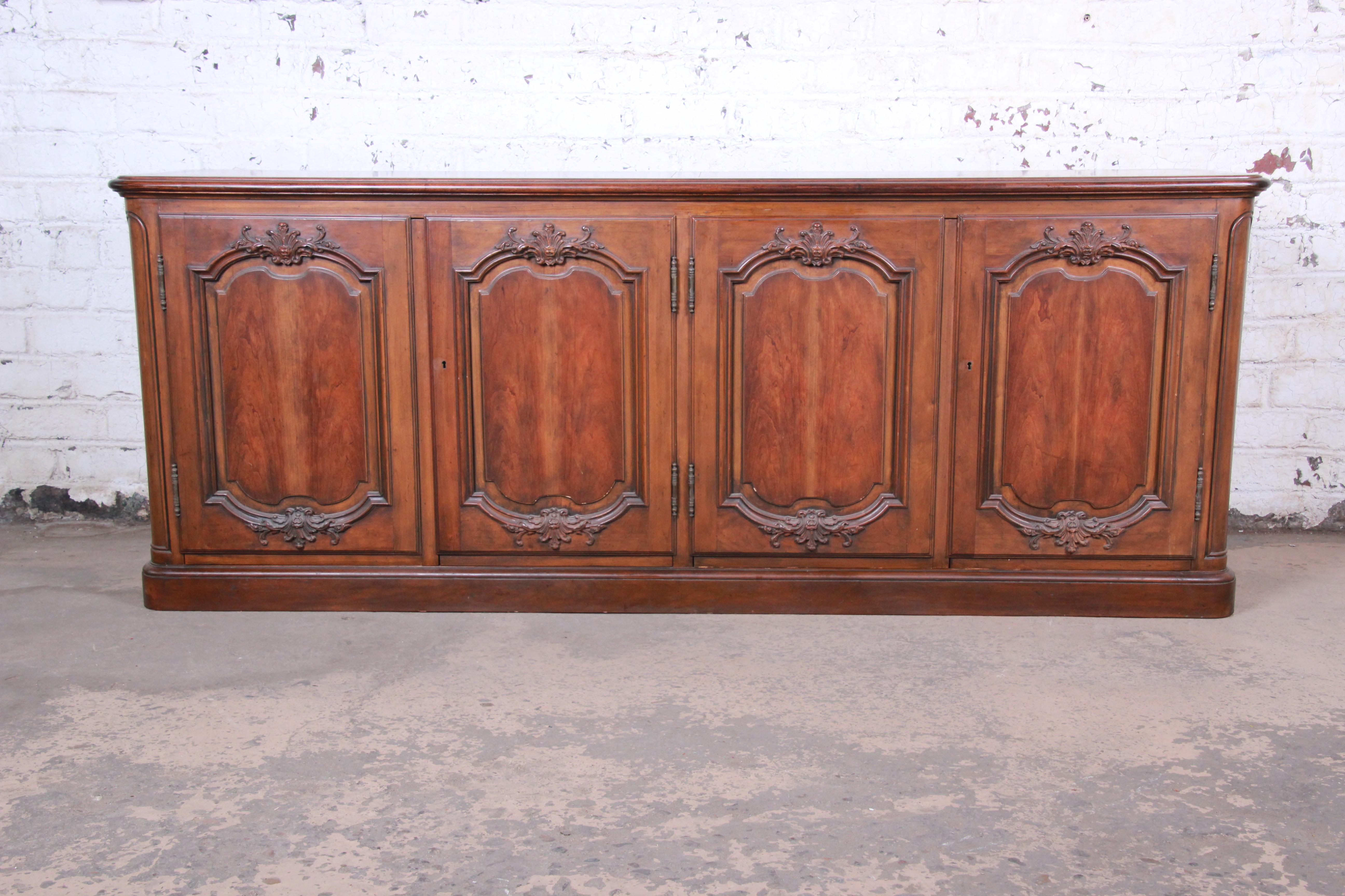 An exceptional French Country carved cherry wood sideboard or bar cabinet by Baker Furniture. The sideboard features gorgeous cherry wood grain with recessed panel doors and floral carvings. It offers ample storage, with two dovetailed drawers and