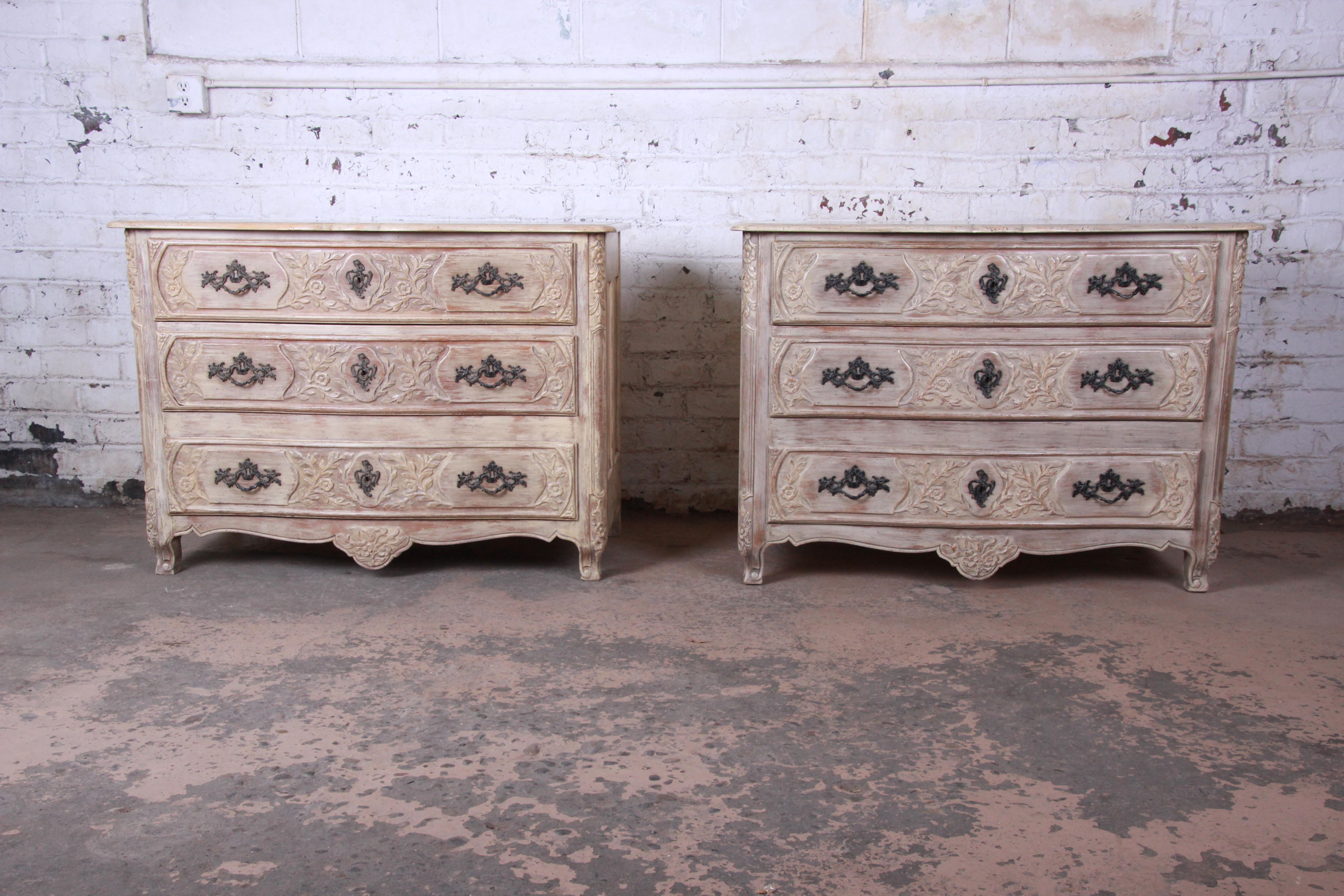 An outstanding pair of French Provincial bachelor chests or large bedside tables by Baker Furniture. The chests feature gorgeous French carved details and the original white-washed finish. They offer ample storage, each with three deep dovetailed