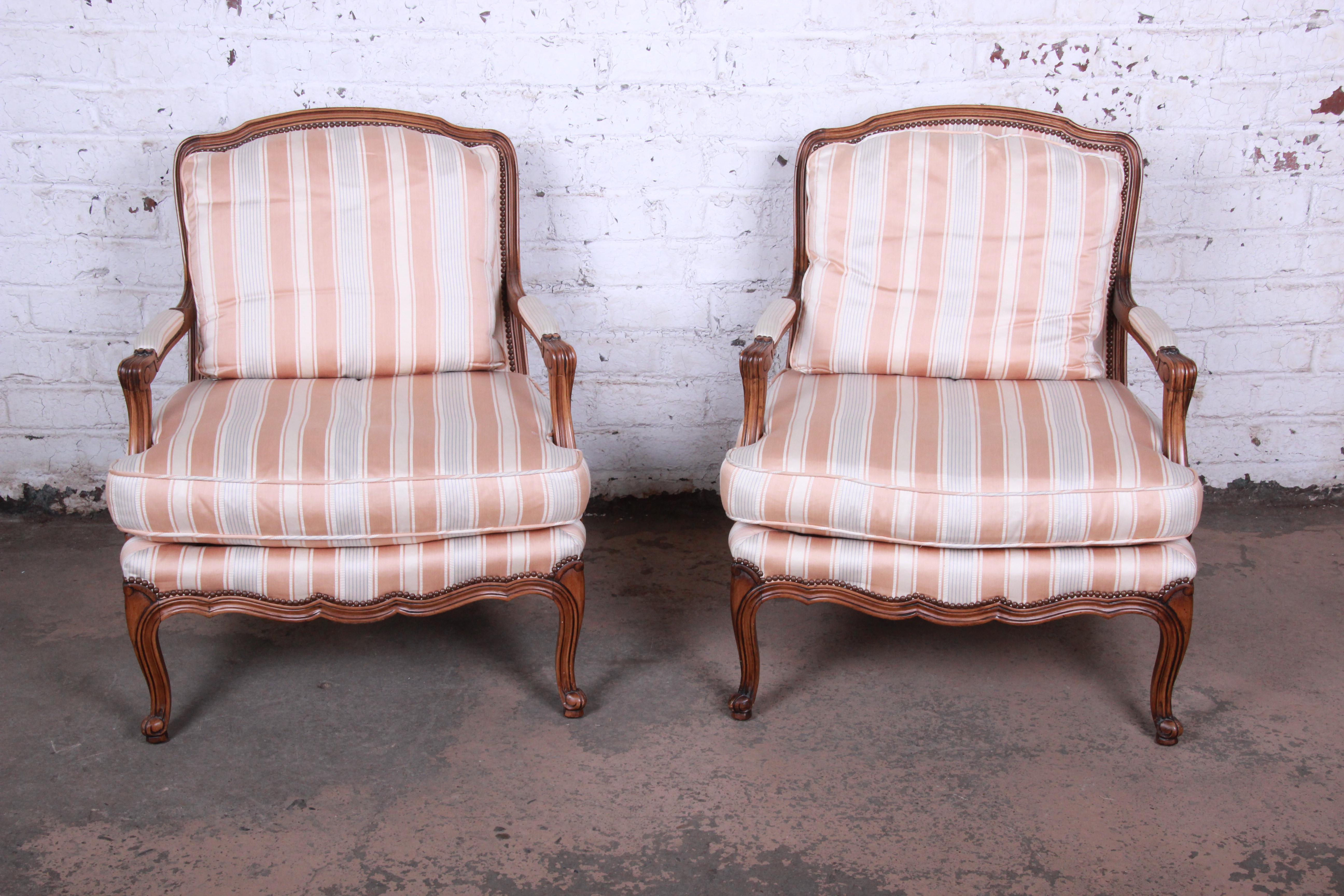 An exceptional pair of open arm bergere chairs in the French Provincial Louis XV style by Baker Furniture. The chairs feature beautifully carved solid walnut frames, original striped upholstery in light pink, light blue, and ivory, and comfortable