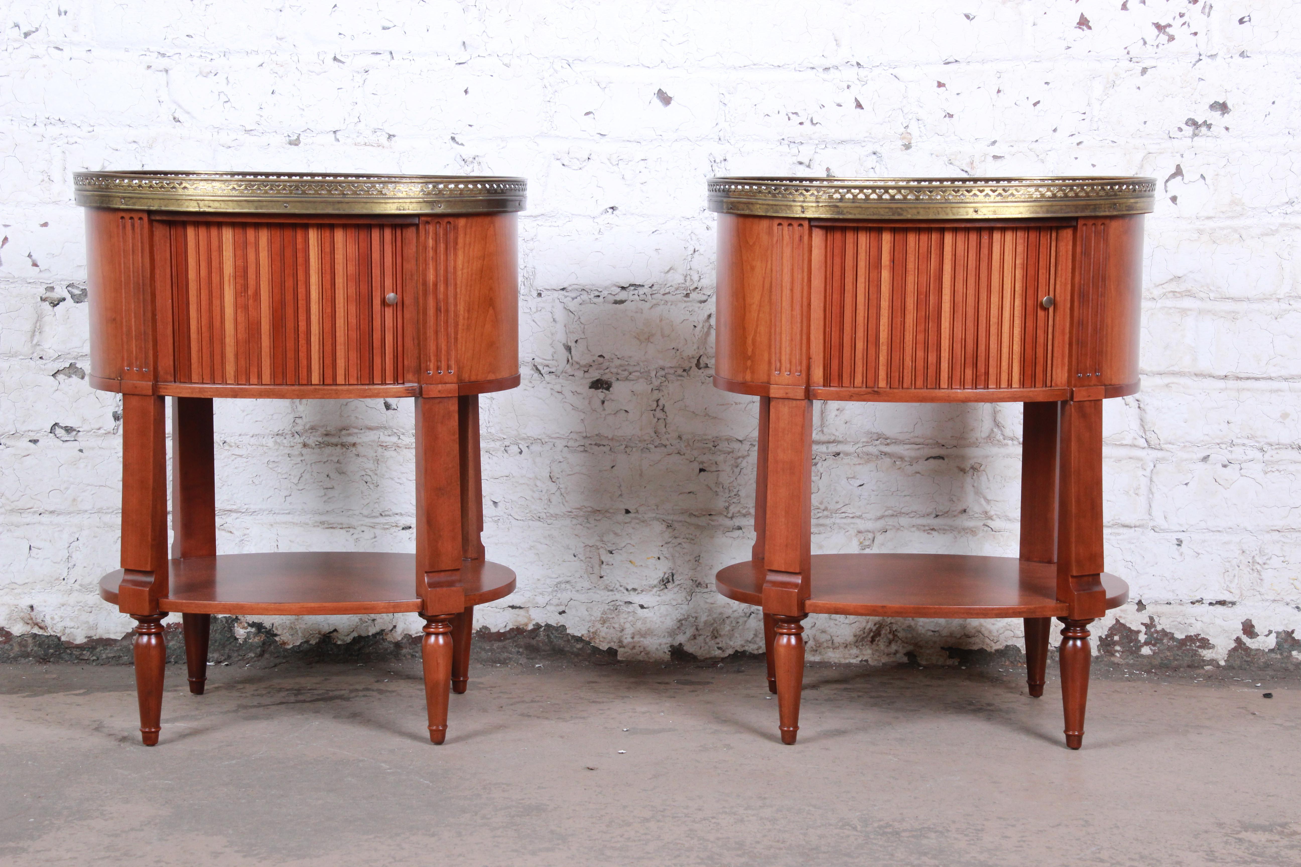 An exceptional pair of French Regency Louis XVI nightstands or end tables by Baker Furniture. The nightstands feature gorgeous cherry wood grain and a pierced brass gallery. They each have cabinet space behind a tambour door and a lower shelf. The