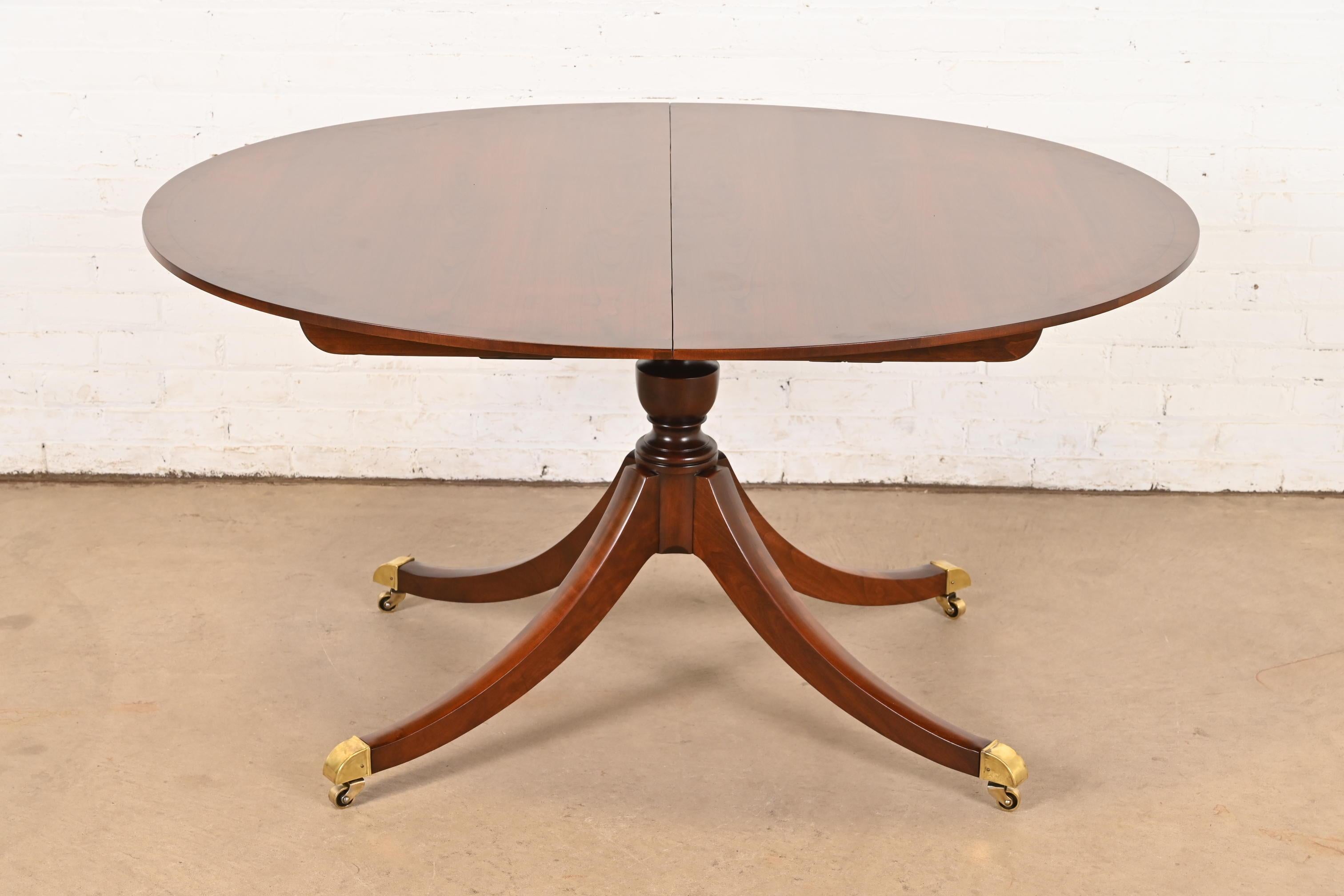Exceptionnelle table de salle à manger à pied de style géorgien ou régence.

Par Baker Furniture

USA, Circa 1980

Magnifique plateau en merisier à bandes assorties, avec piédestaux sculptés, et pieds coiffés de laiton sur roulettes en