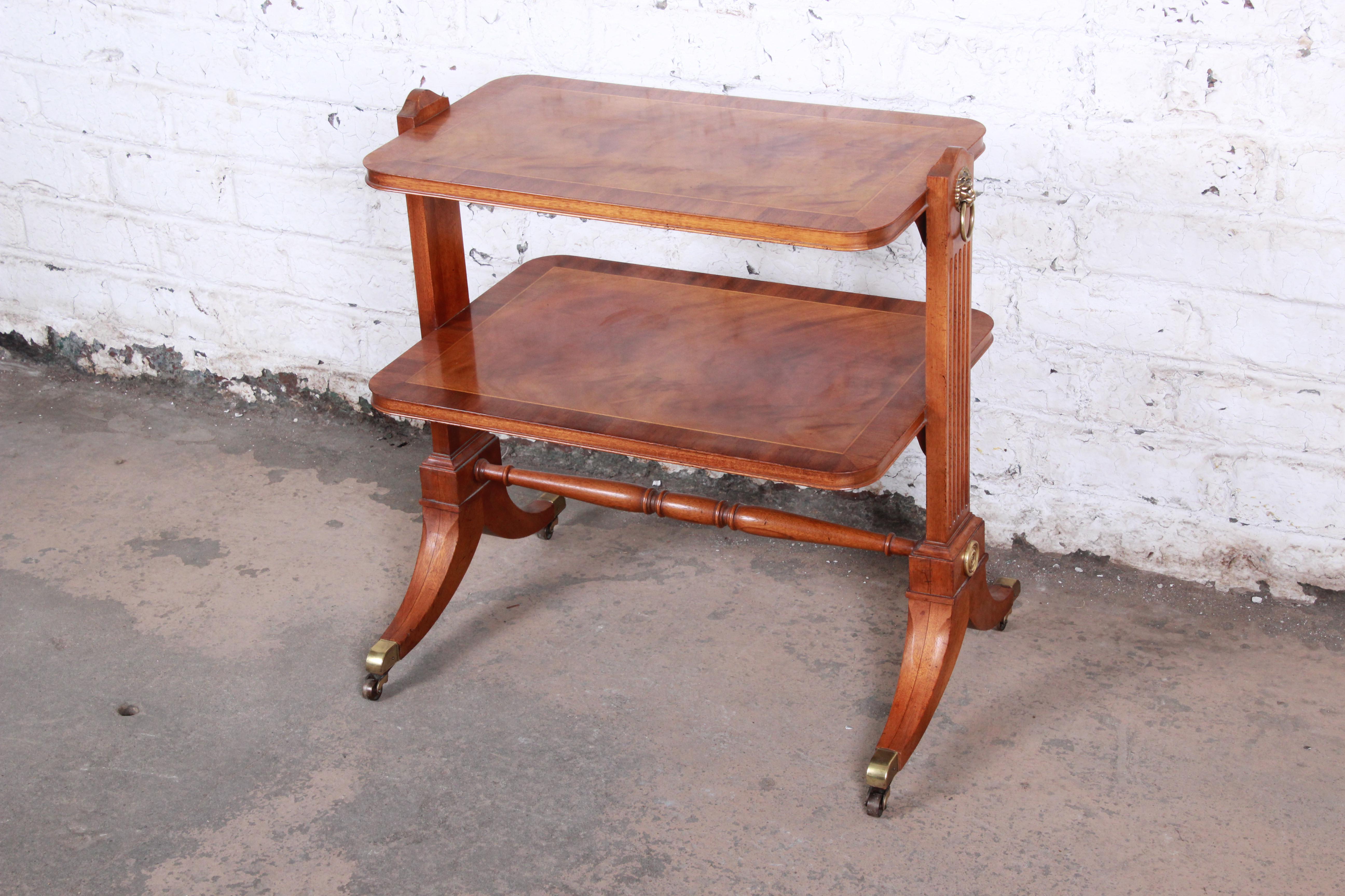 An exceptional Georgian banded mahogany two-tier occasional table by Baker Furniture. The table features gorgeous flame mahogany wood grain with nice carved details and brass accents, including brass-tipped feet and casters. The original baker label