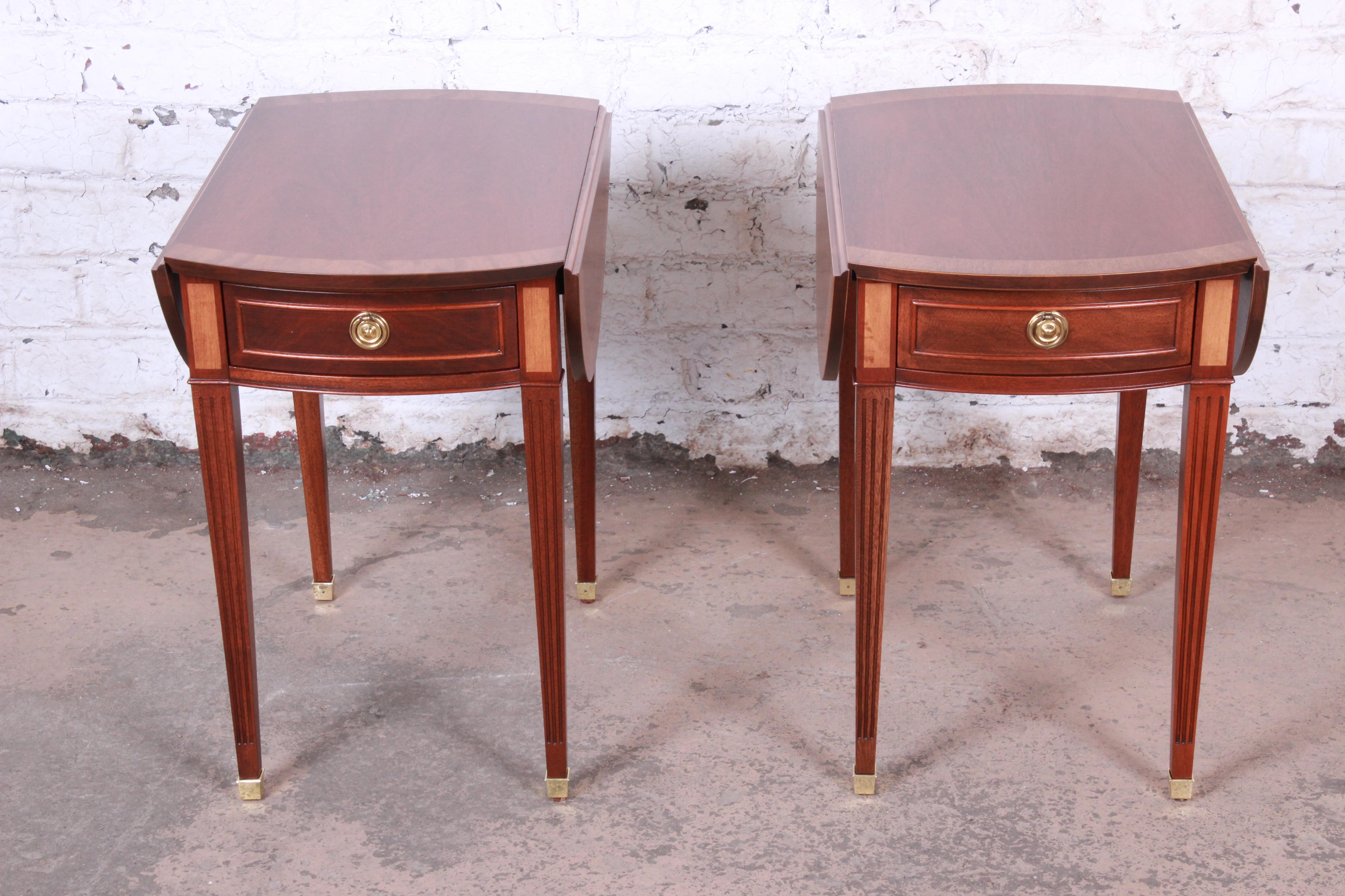 An exceptional pair of Georgian style banded mahogany drop-leaf pembroke side tables by Baker Furniture. The tables feature gorgeous mahogany wood grain with a banded edge and beautiful inlaid details. They offer good storage, each with a single