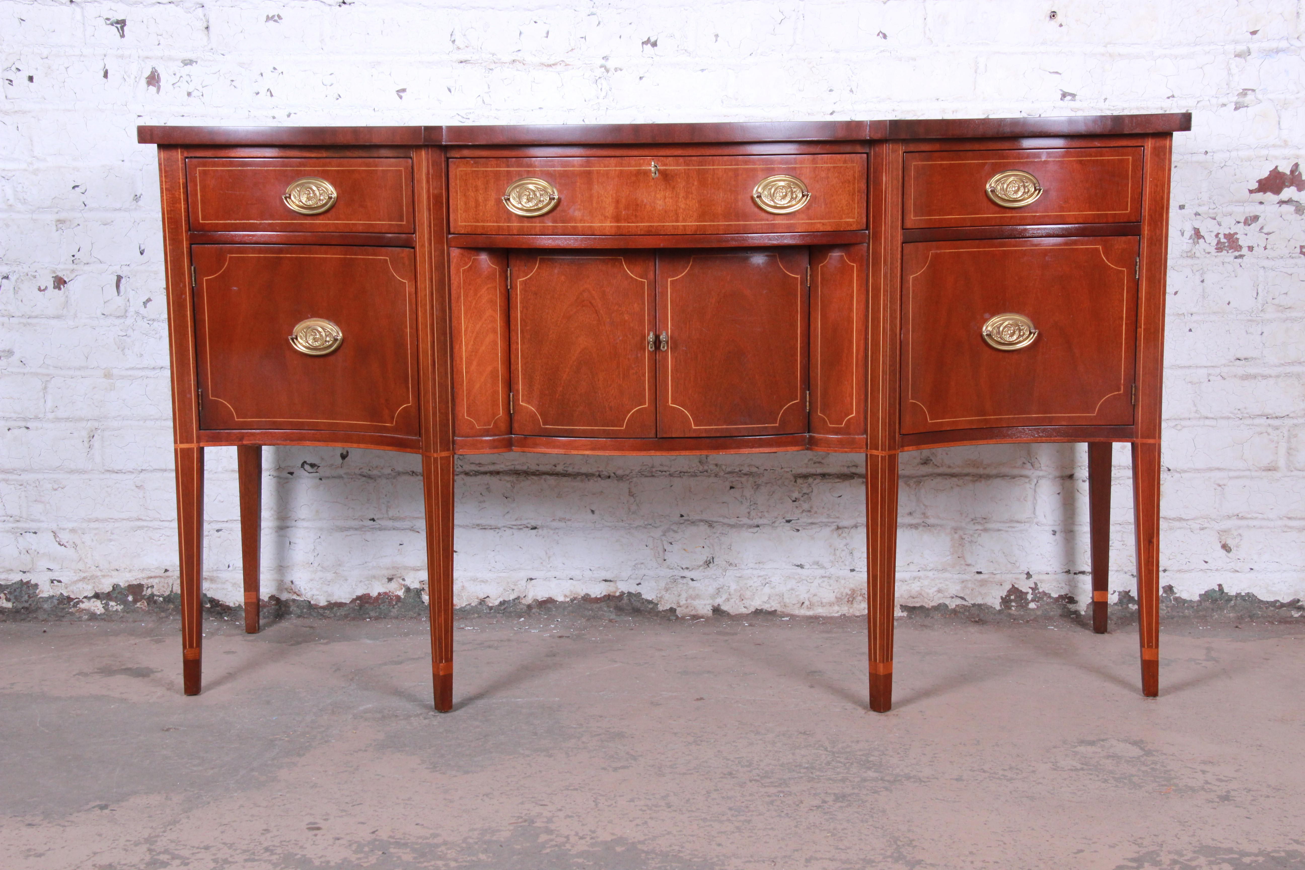 A gorgeous Hepplewhite style mahogany sideboard buffet by Baker Furniture. The sideboard features stunning mahogany wood grain with string inlay and a nice bow front design. The case rests on tall tapered legs. It offers ample storage, with three