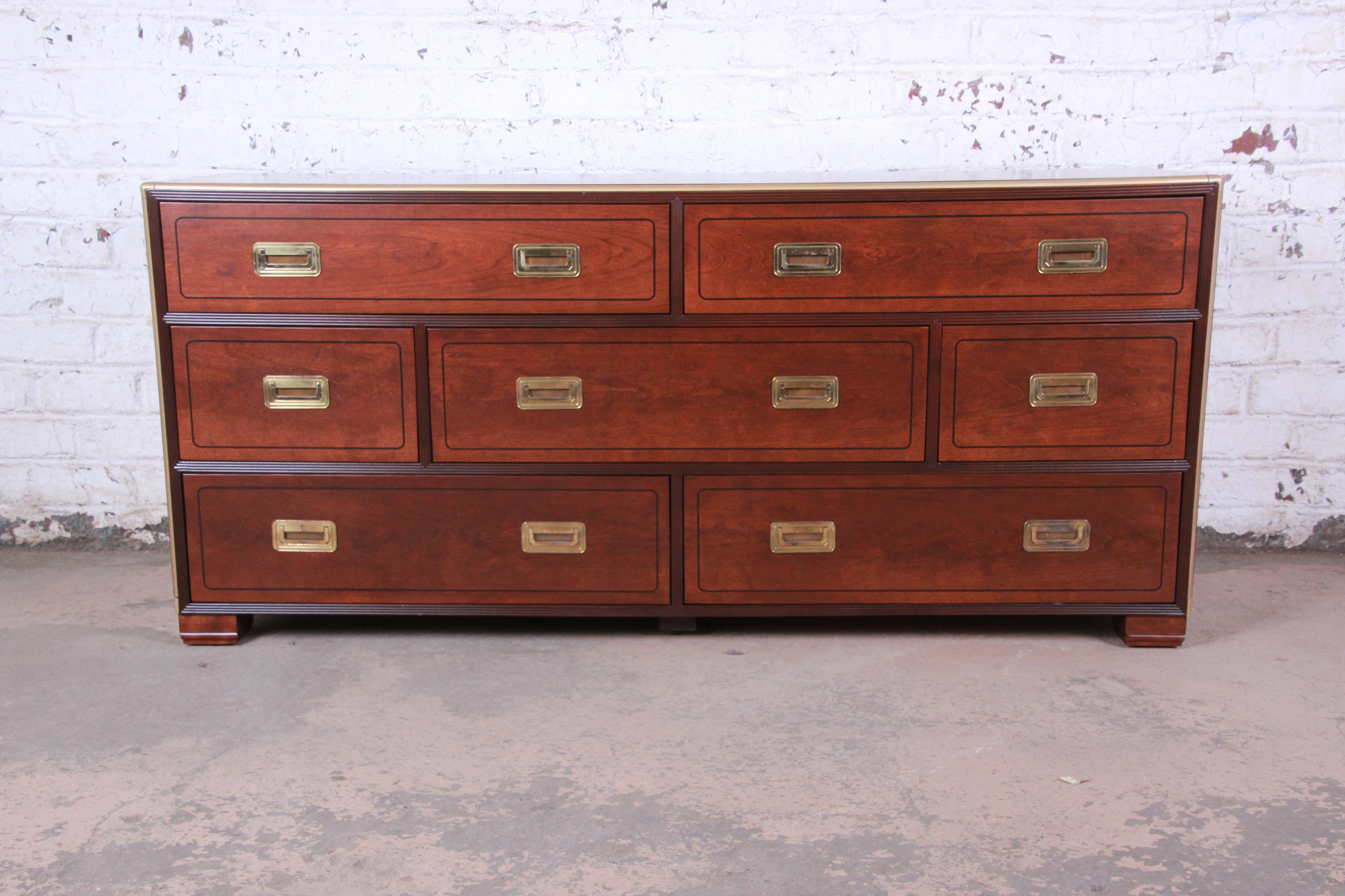 An exceptional Hollywood Regency Campaign style long dresser or credenza by Baker Furniture. The dresser features gorgeous walnut wood grain with inlaid details and brass trim. It offers ample storage, with seven deep dovetailed drawers. Hardware is
