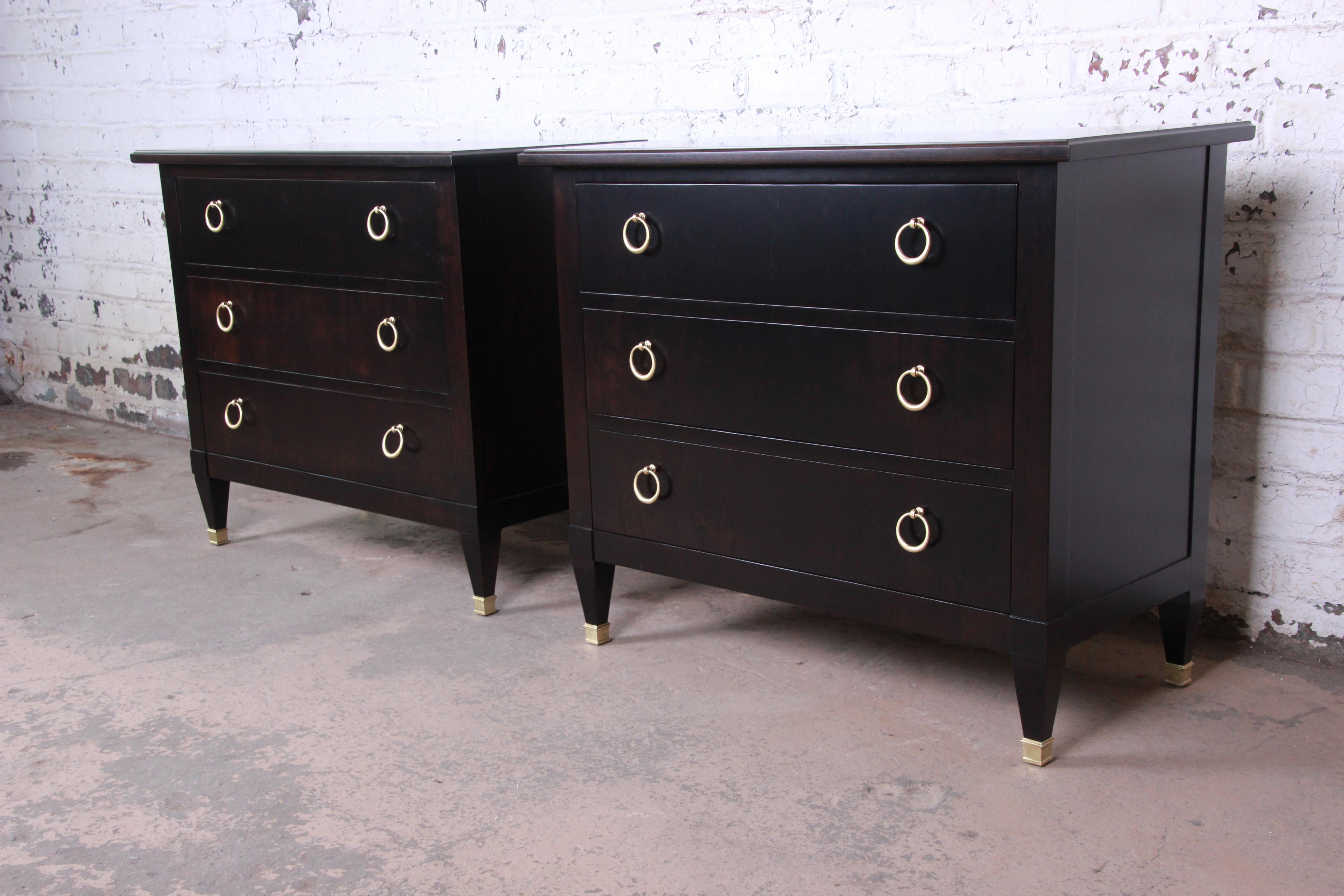 A gorgeous pair of Hollywood Regency large bedside tables or bachelor chests by Baker Furniture. The chests feature stunning walnut wood grain with a newly ebonized finish and original polished brass hardware and brass-tipped feet. They offer ample