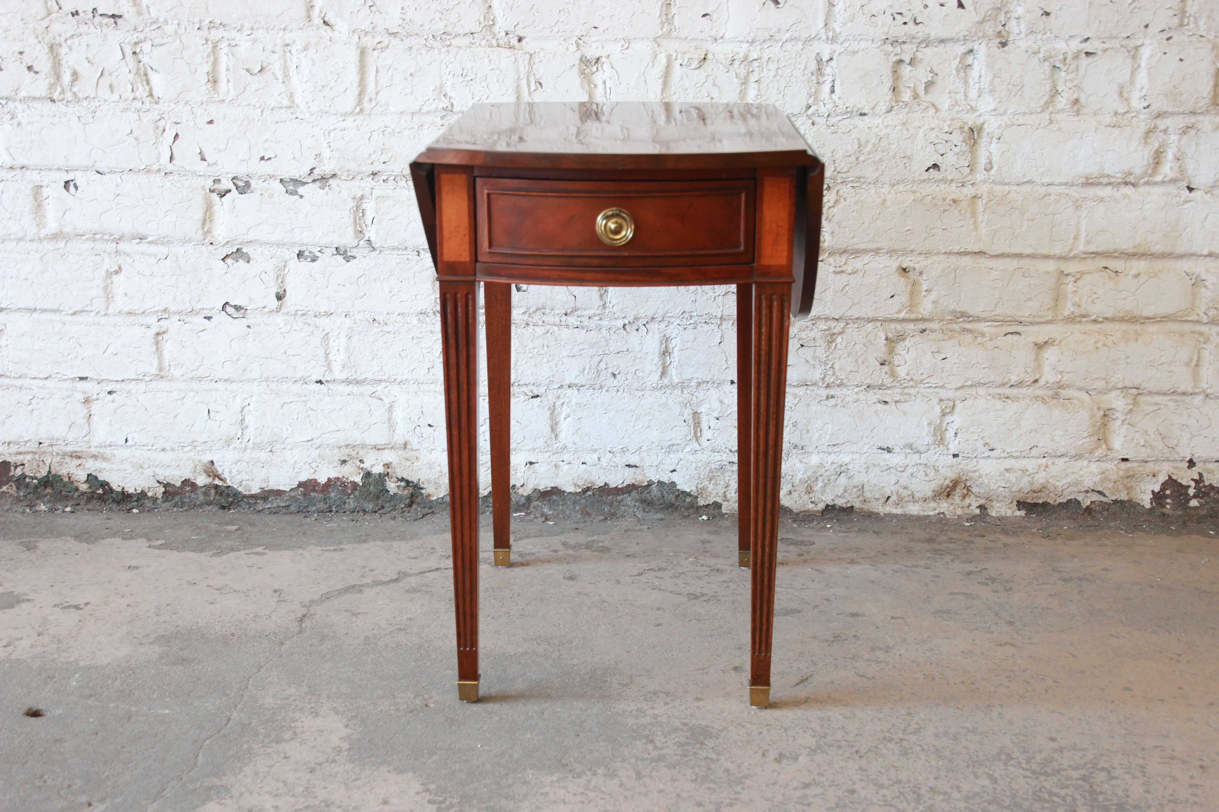 Offering a very nice mahogany and satinwood inlaid drop-leaf side table by Baker Furniture. The table has tapered legs with brass details at the feet. The table has a smooth sliding drawer and round brass pull. Each side has a drop-leaf that