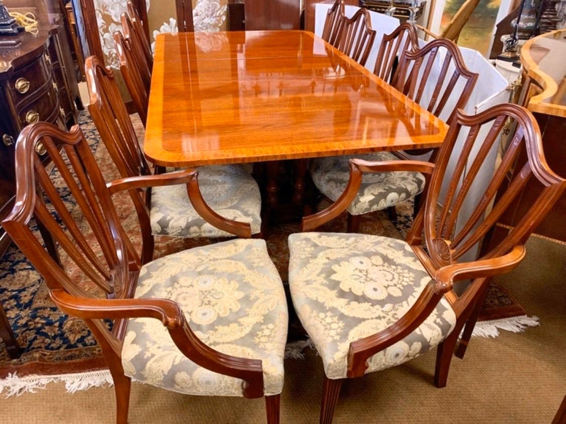 Stunning Baker Furniture dining room set includes a double pedestal mahogany table with one leaf and ten shield-back chairs which include two arms and eight side chairs. Table features a banded inlay all around and brass paw feet. All Baker