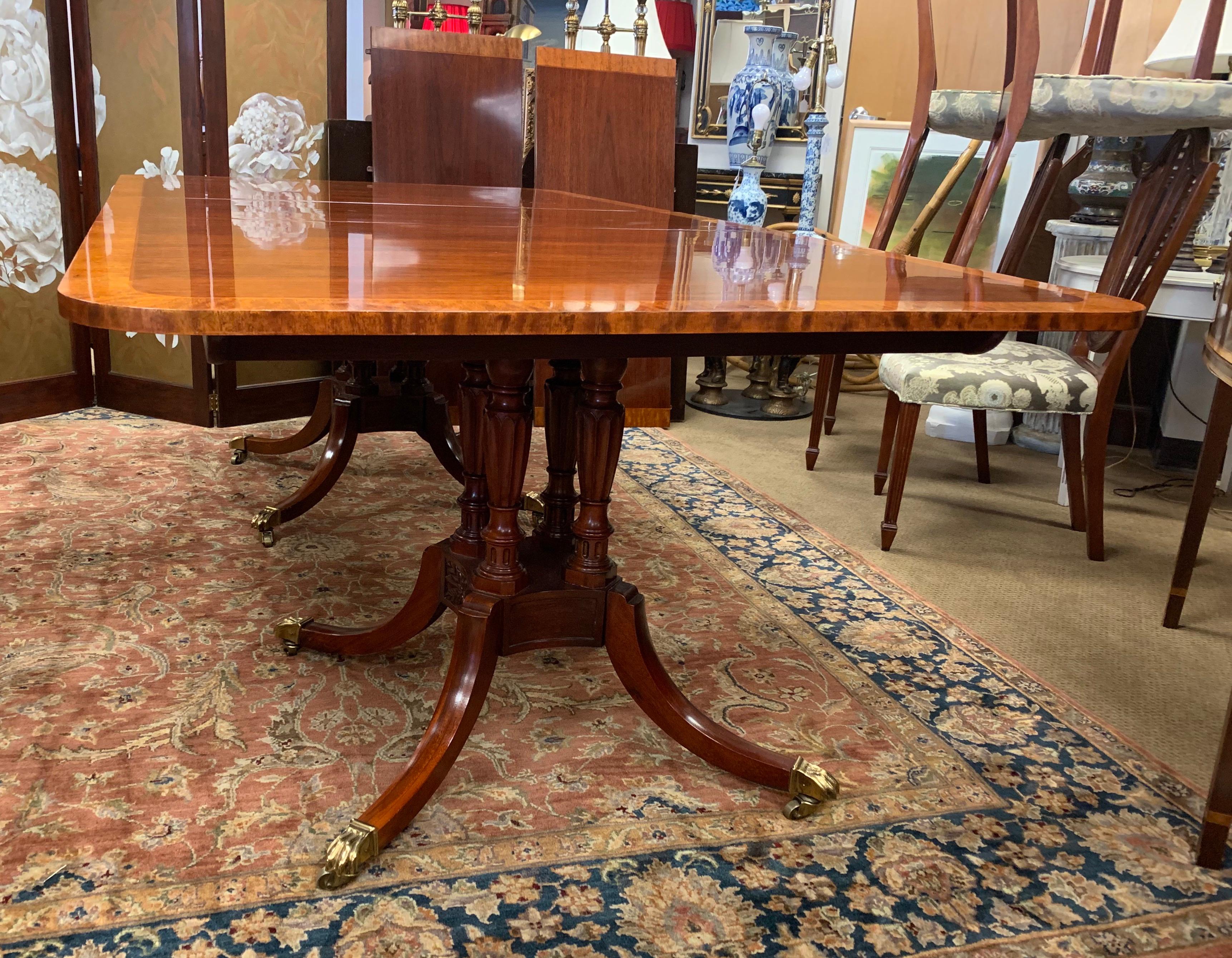 Stunning Baker Furniture mahogany double pedestal dining table features a banded satinwood inlay all around and four turned columns that are supported by a square base with four saber legs that curve outward with brass feet on casters. Comes with