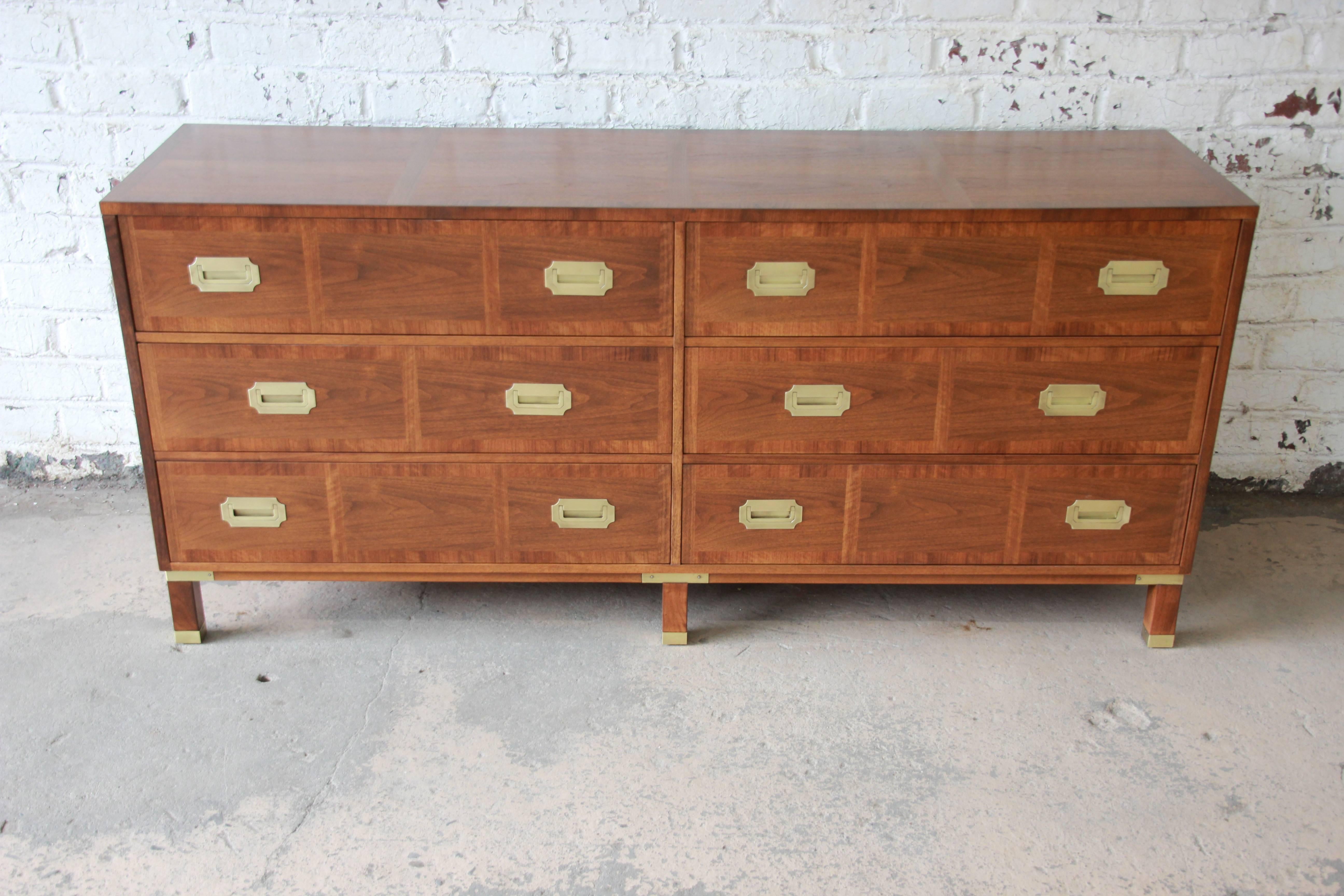A stunning newly refinished midcentury Hollywood Regency Campaign style dresser or credenza from the Milling Road line by Baker Furniture. The dresser features rich walnut wood grain with a banded edge and beautiful polished brass hardware and