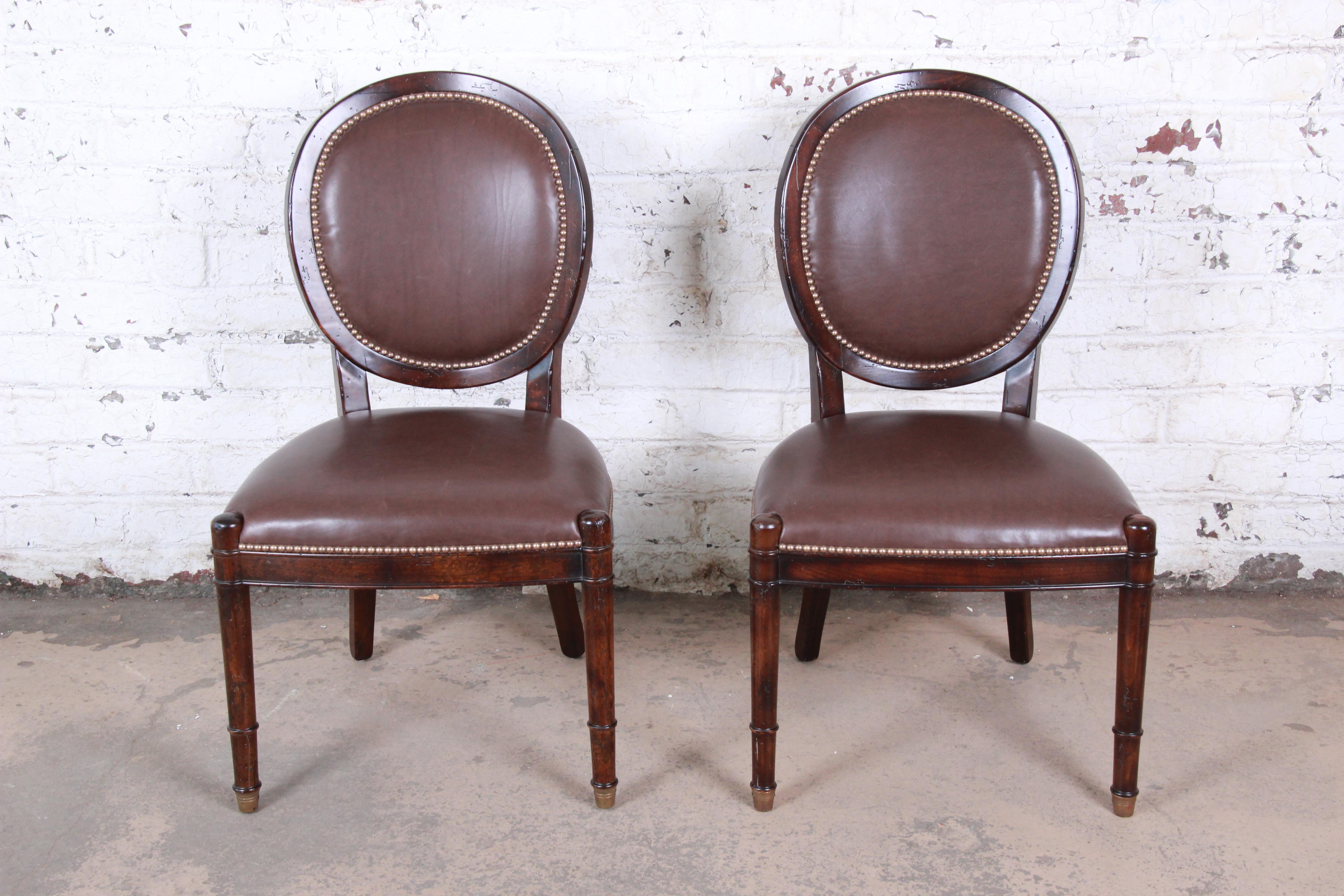 A gorgeous pair of balloon back side chairs from the Milling Road line by Baker Furniture. The chairs features solid walnut frames with brass-tipped feet and high-end studded brown leather upholstery. The original labels are present on the underside